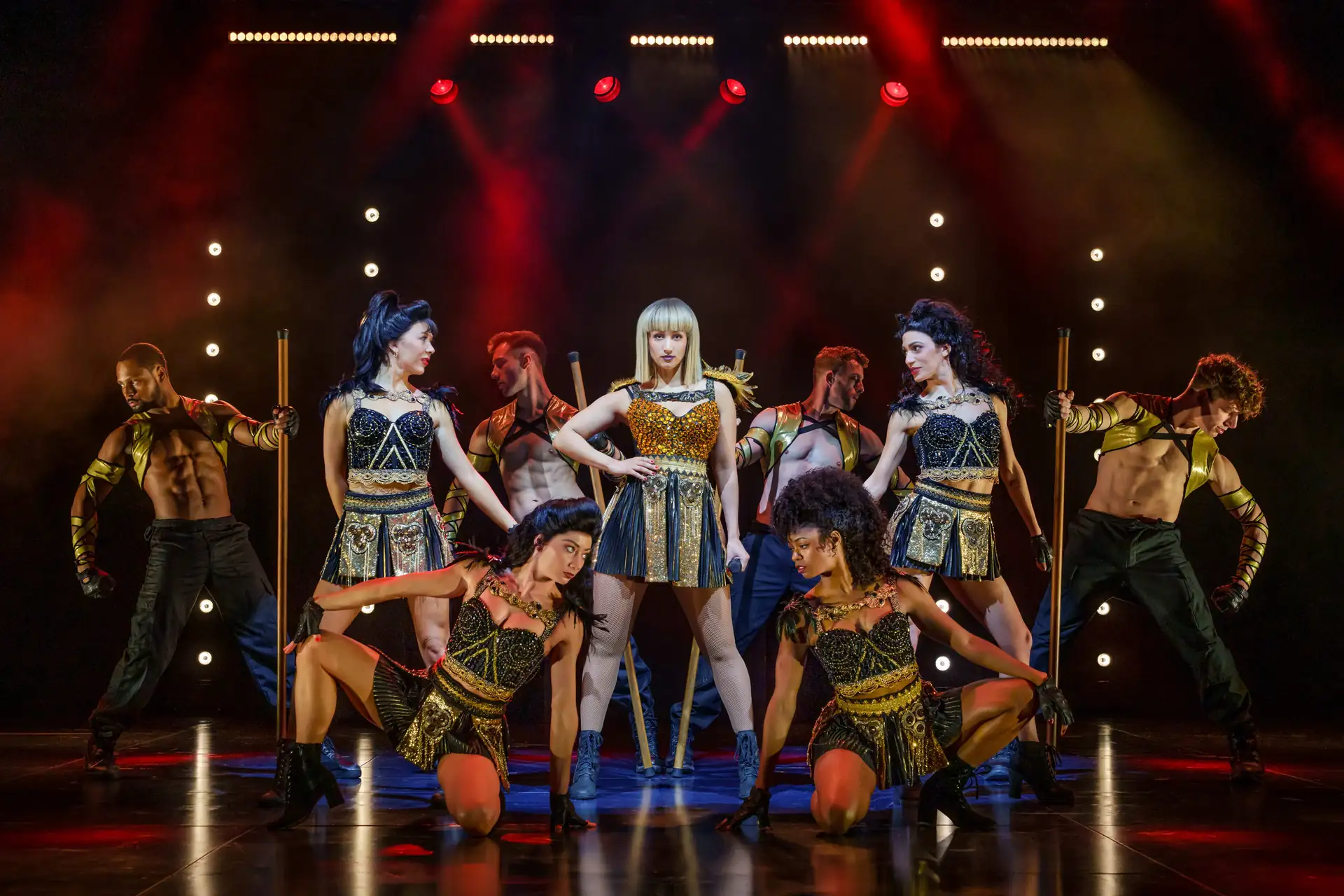 The Bodyguard – The Musical Tour: Acht Tänzer in gold-schwarzen Kostümen posieren auf einer Bühne mit roter Beleuchtung, die zentrale Tänzerin trägt ein goldfarbenes Oberteil und schwarzen Rock.