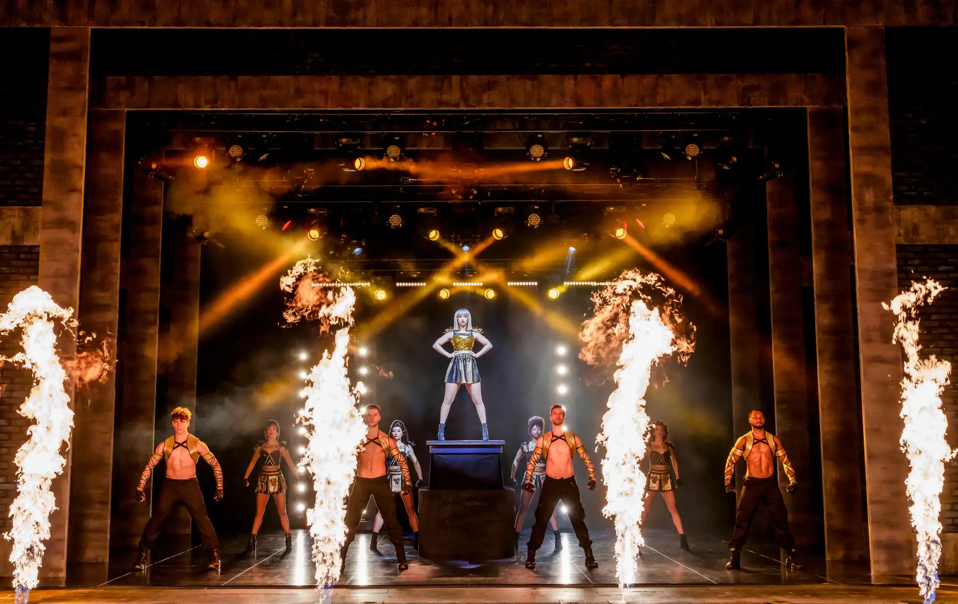 The Bodyguard – The Musical Tour: Eine Frau steht auf einer erhöhten Plattform in der Mitte einer Bühne, umgeben von sechs Tänzern in schwarzen Outfits und vier großen Flammenfontänen.