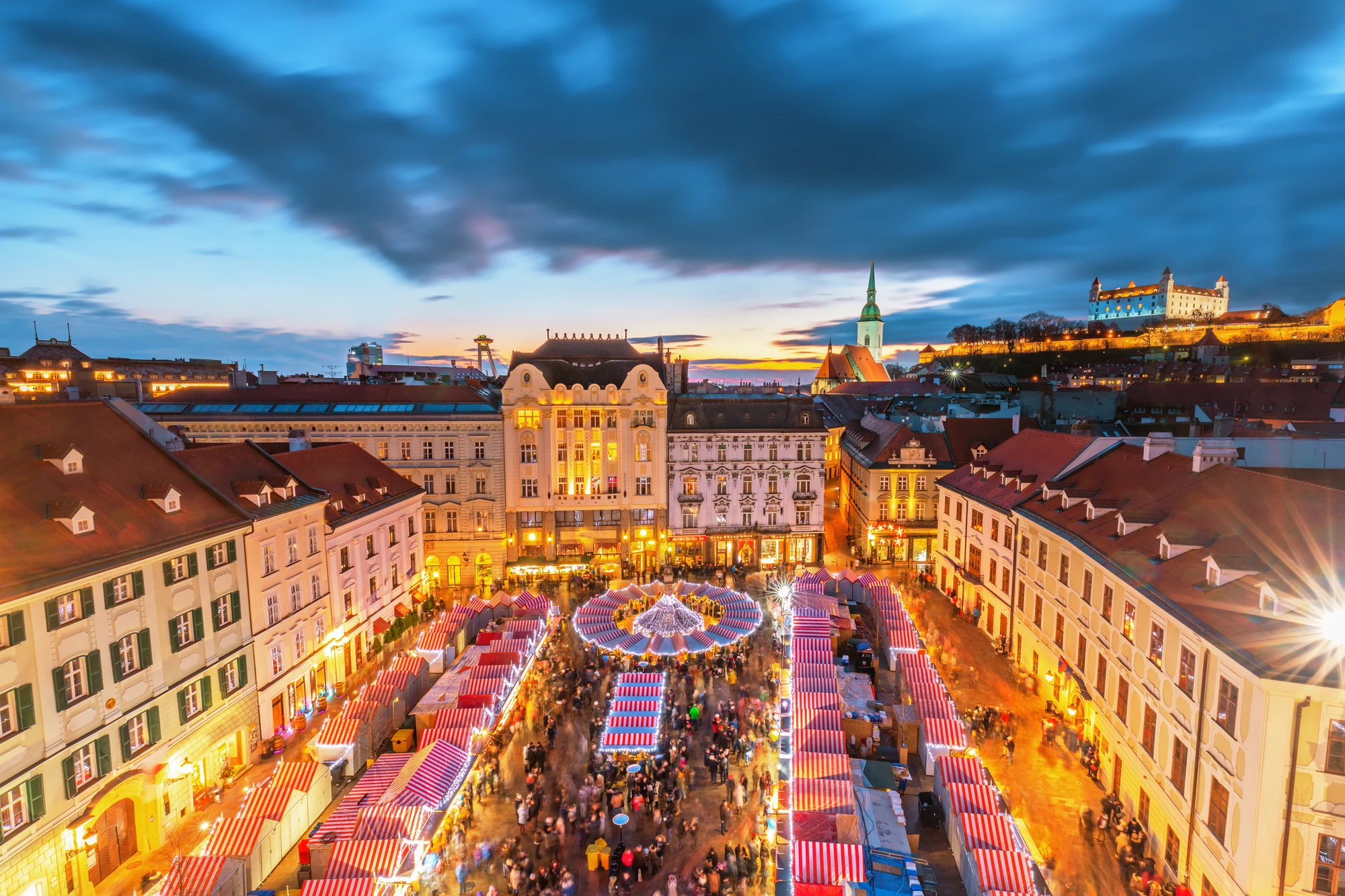 Weihnachtsmarkt mit vielen Ständen und Menschen auf dem Hauptplatz in der historischen Altstadt von Bratislava bei Abenddämmerung, im Hintergrund Burg und Kirchturm