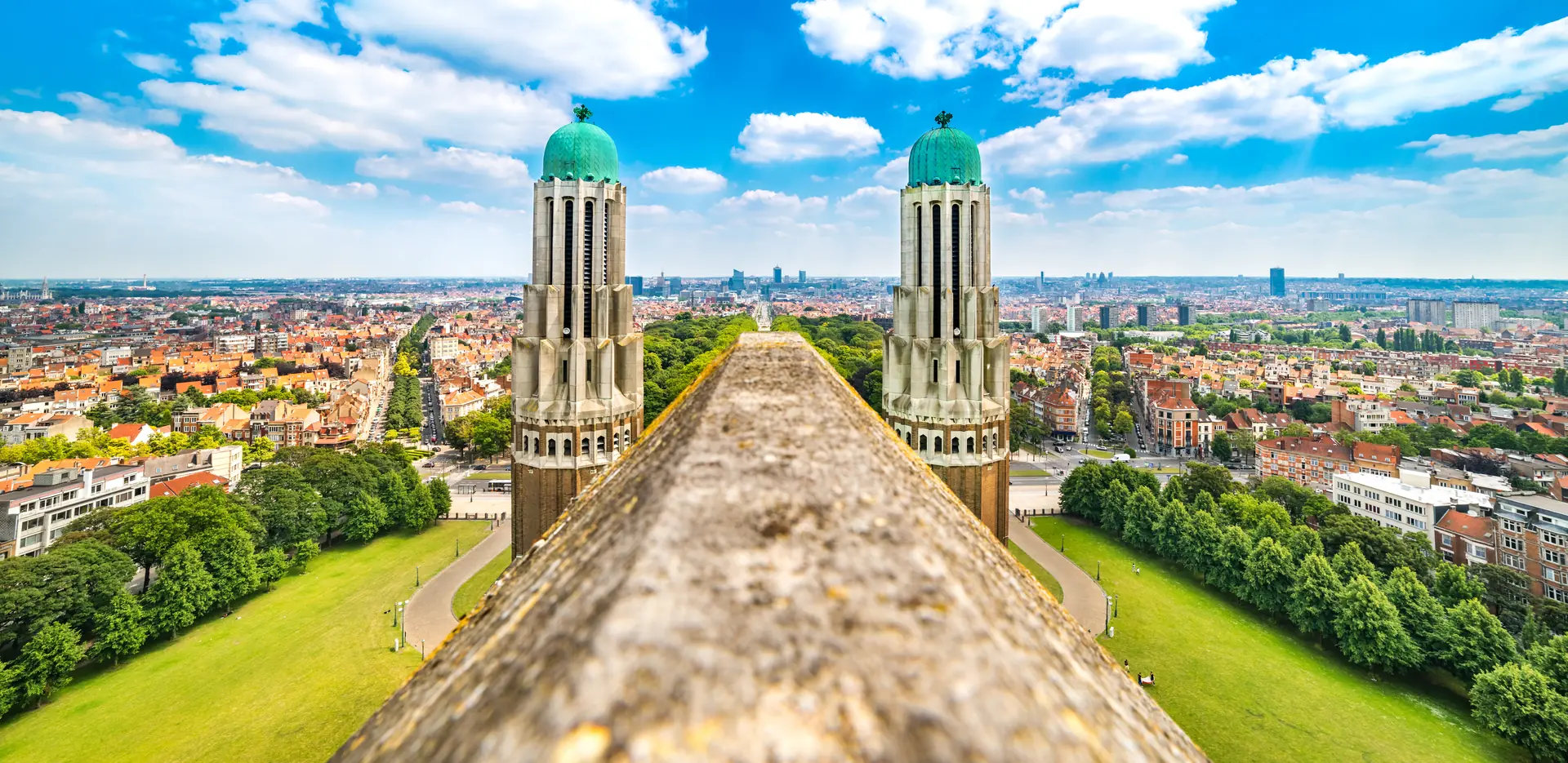 Von der Dachspitze der Basilika des Heiligen Herzens mit traumhaftem Blick auf Brüssel. Der sonnige Ausblick zeigt die Stadt und die umliegenden Grünflächen, flankiert von den markanten Türmen der Basilika auf der linken und rechten Seite.