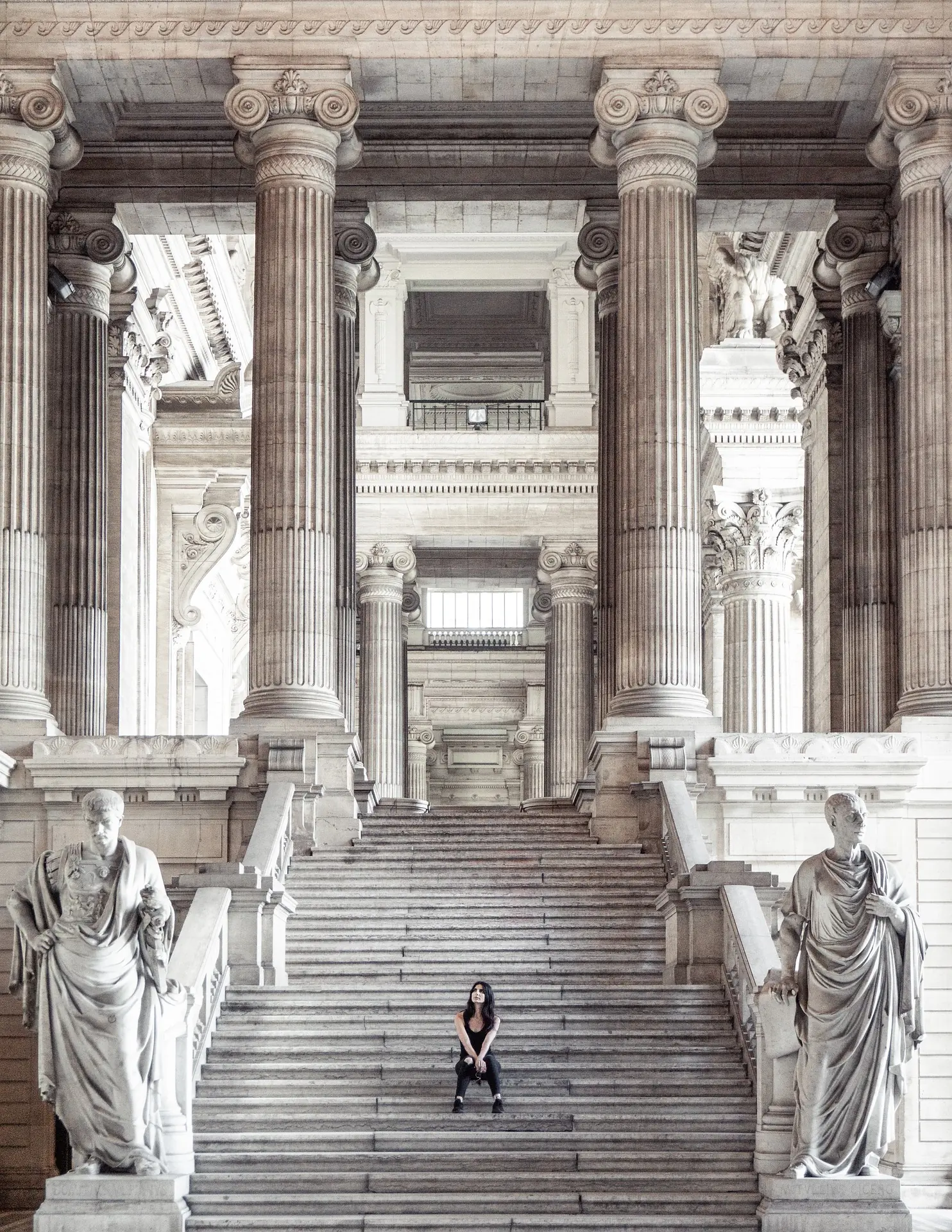 Der Justizpalast in Brüssel bei Tageslicht, mit seiner imposanten Fassade. Eine Frau sitzt auf dem Treppenaufgang, was die Größe des Gebäudes zusätzlich betont.