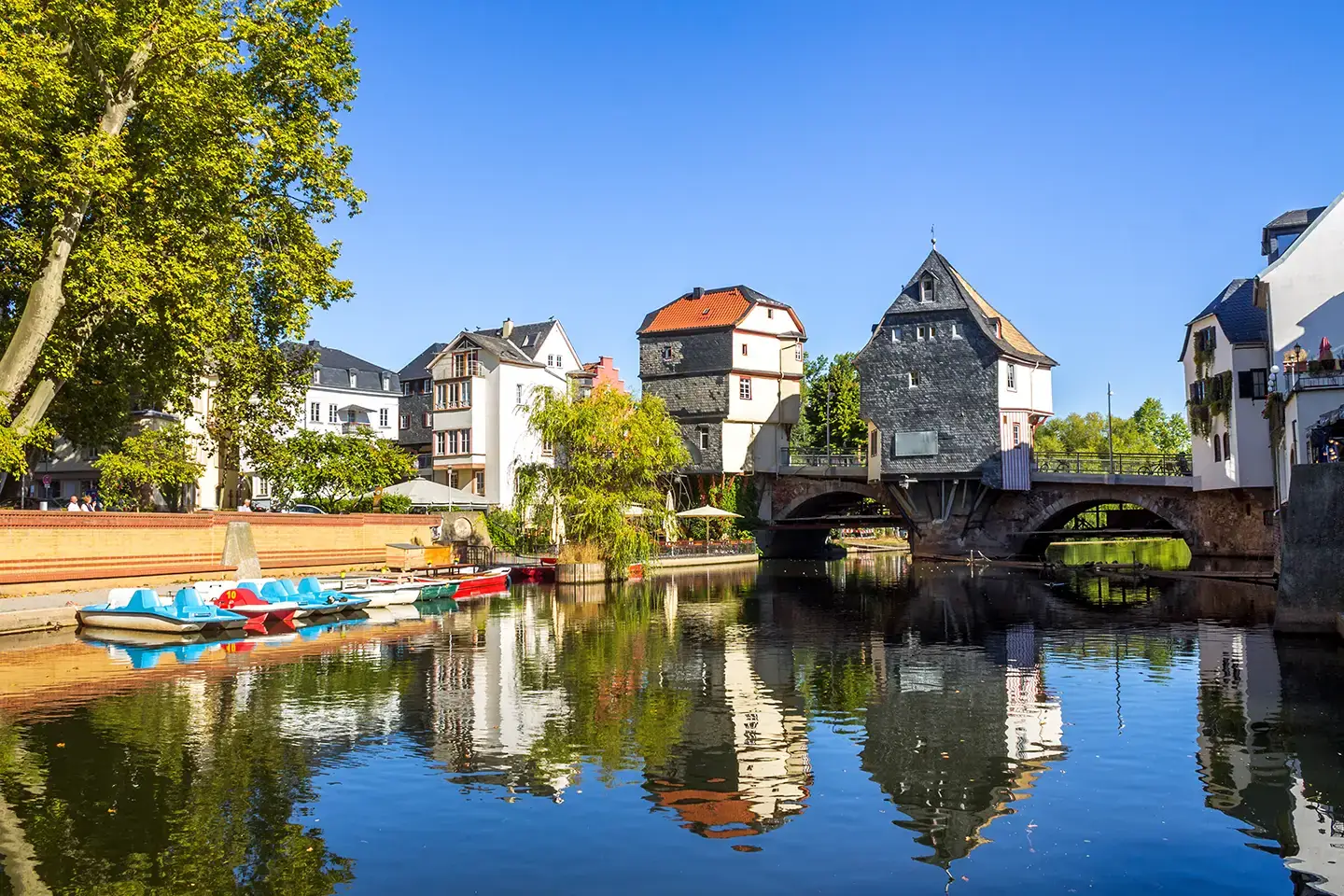 Stadtmotiv Bad Kreuznach mit geparkten Tretboten auf dem Fluss Nahe vor einer kleinen historischen Brücke