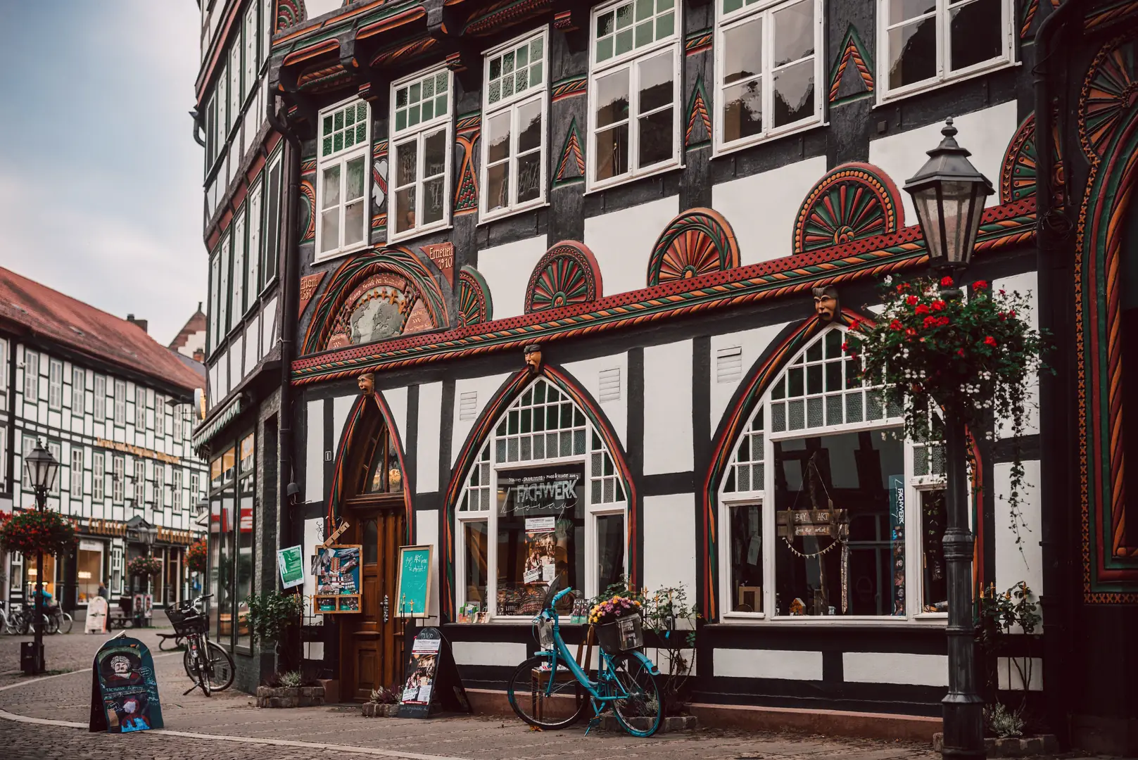 Prächtiges Fachwerkhaus in der Altstadt von Einbeck mit kunstvoll verzierten Fassadenelementen in traditionellen Farben. Die reich dekorierten Balken und filigranen Fensterbögen verleihen dem historischen Gebäude einen einzigartigen Charme. Im Vordergrund ein blaues Fahrrad und blühende Blumen, die das malerische Stadtbild unterstreichen.