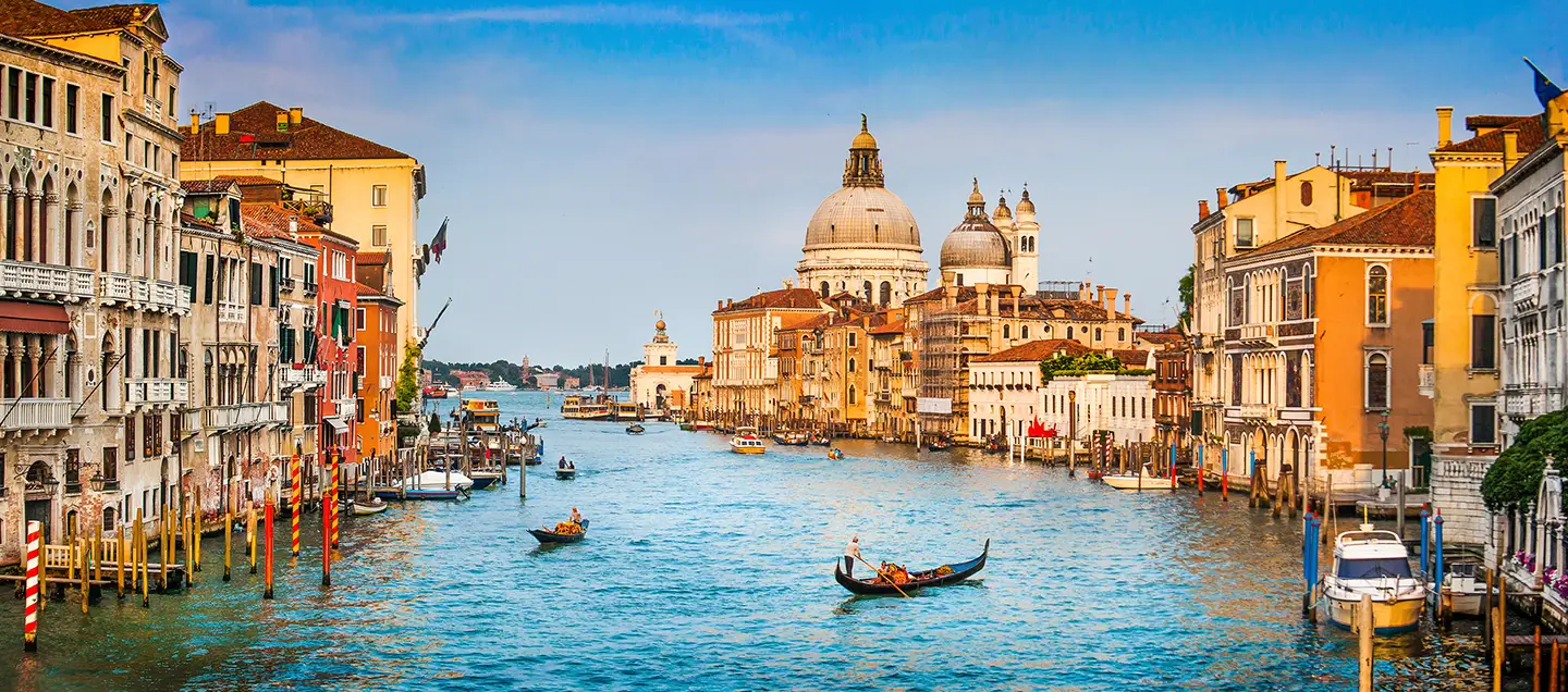 Ein Blick auf den Canal Grande in Venedig bei Tageslicht zeigt angelegte und fahrende Gondeln und Boote. Entlang des Kanals reihen sich farbenfrohe Häuser, die die malerische Kulisse der Stadt unterstreichen.