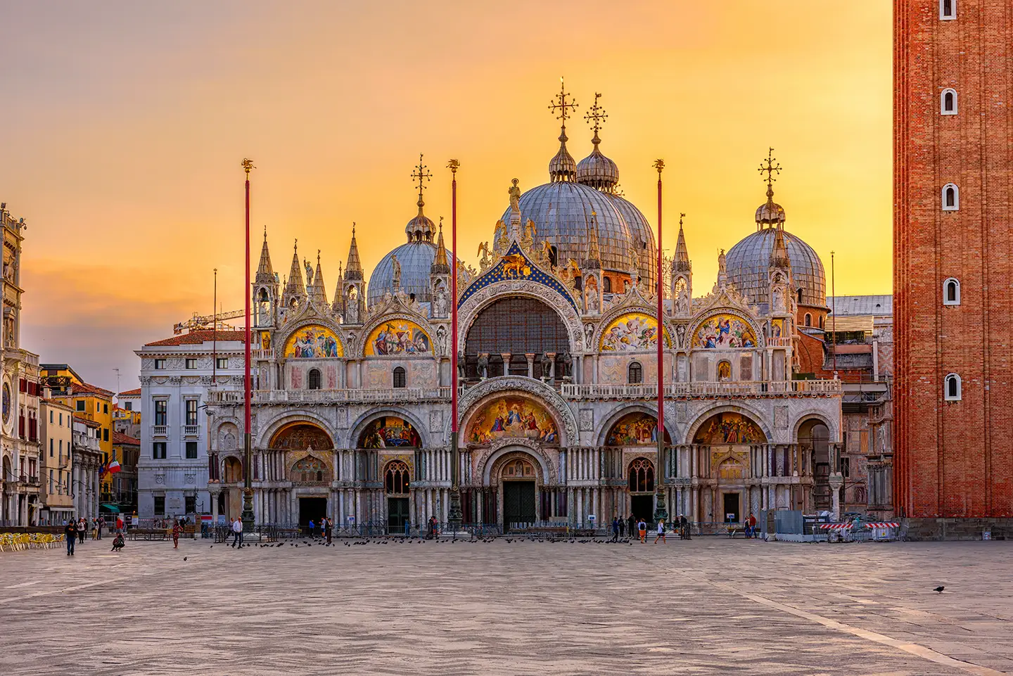 Die Frontansicht des Markusdoms in Venedig erstrahlt im goldenen Licht der Abenddämmerung. Auf dem Platz sind nur wenige Spaziergänger und Touristen zu sehen, während zahlreiche Tauben friedlich auf dem Platz vor dem Dom verweilen und die Atmosphäre des historischen Ortes unterstreichen.