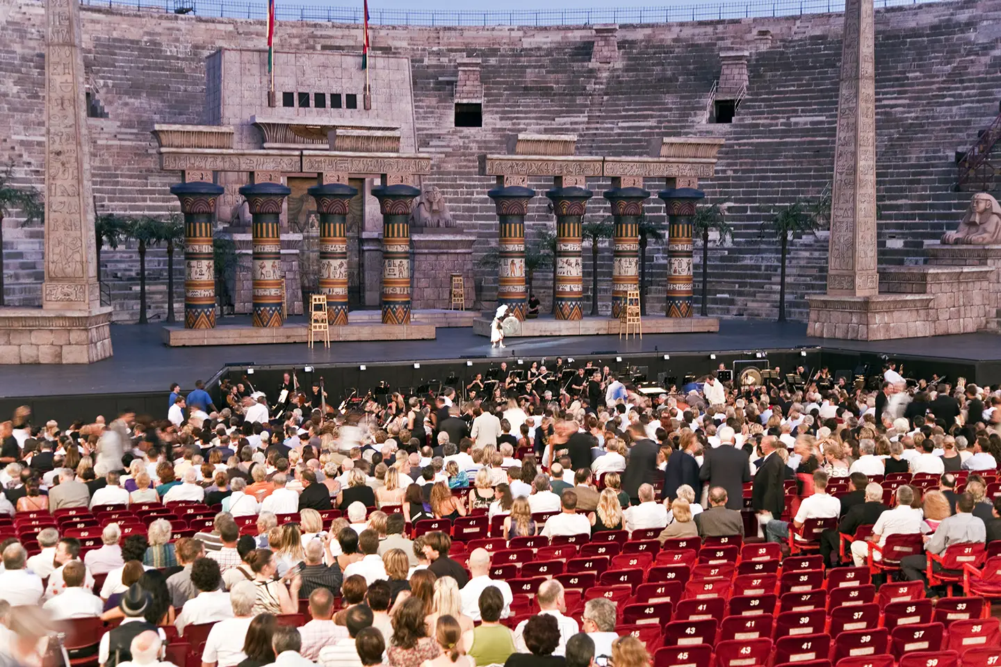 Innenansicht der Arena di Verona, die mit Publikum gefüllt ist. Die beeindruckende Architektur und die Sitzreihen sind deutlich sichtbar, während die Atmosphäre von Vorfreude und Begeisterung geprägt ist.