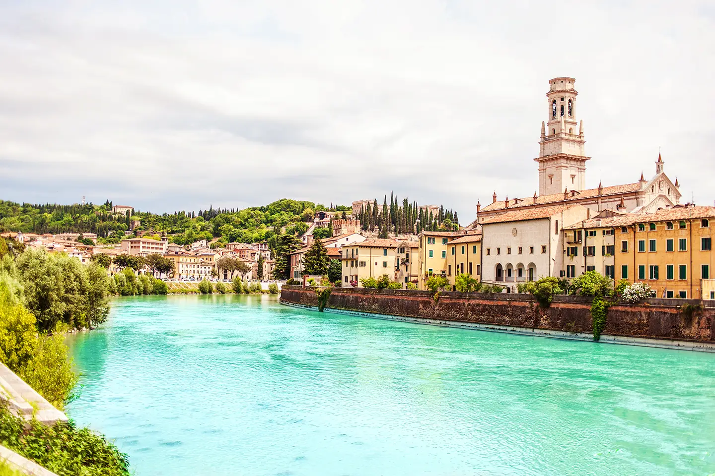 Stadtmotiv von Verona, das den hellen smaragdgrünen Fluss Etsch zeigt, umgeben von weitläufigen grünen Flächen und historischen Gebäuden. Die harmonische Verbindung von Natur und Architektur schafft eine einladende Atmosphäre in der Stadt.
