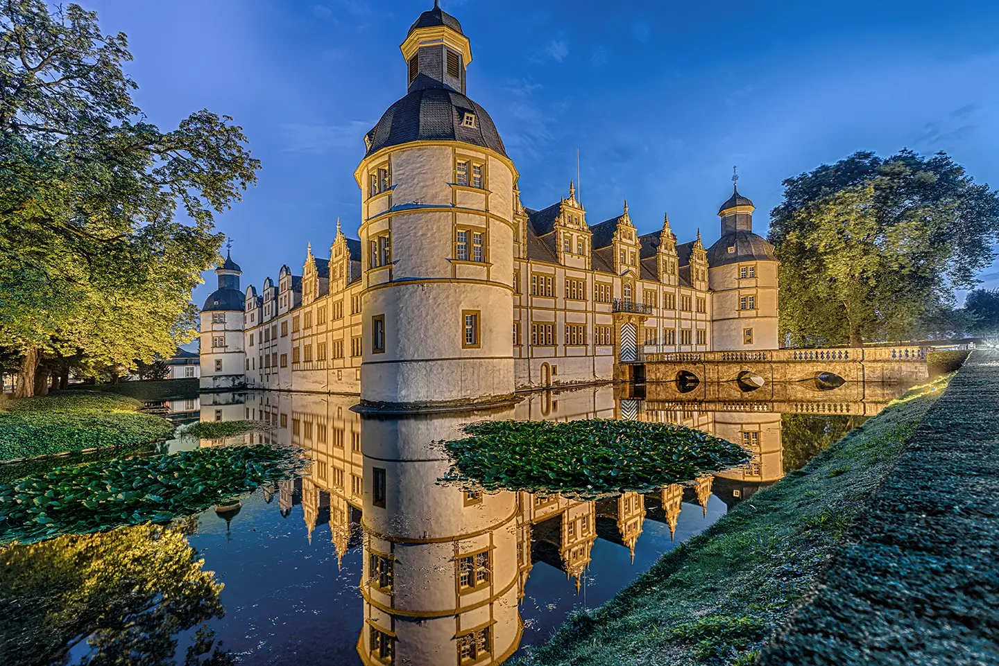 Blick auf das beleuchtete Residenz- und Wasserschloss Neuhaus, umgeben von einem mit Wassergraben mit Seerosen