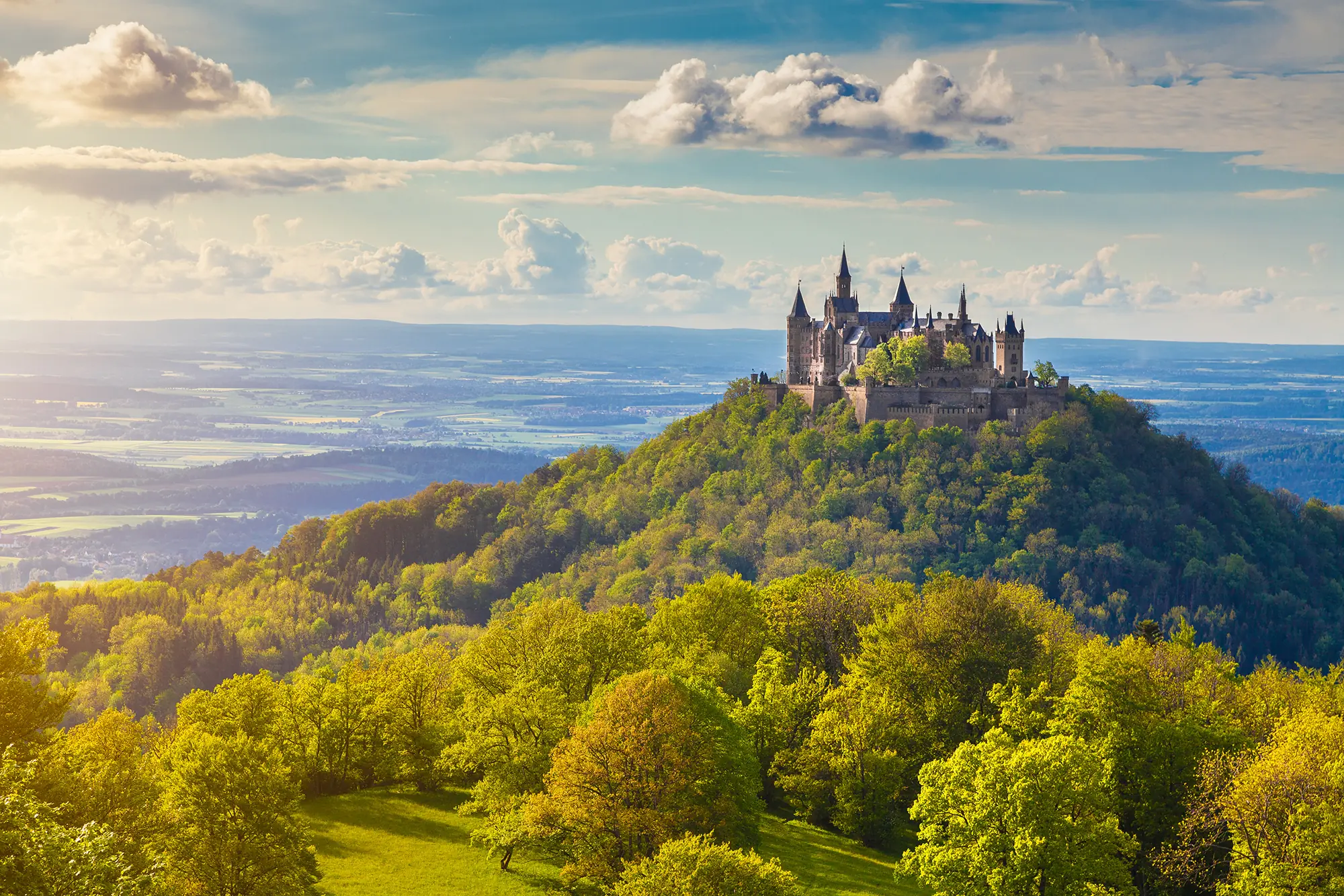 Panoramablick auf Burg Hohenzollern, die majestätisch auf einer grün bewaldeten Anhöhe thront, mit der weiten schwäbischen Alb im Hintergrund.