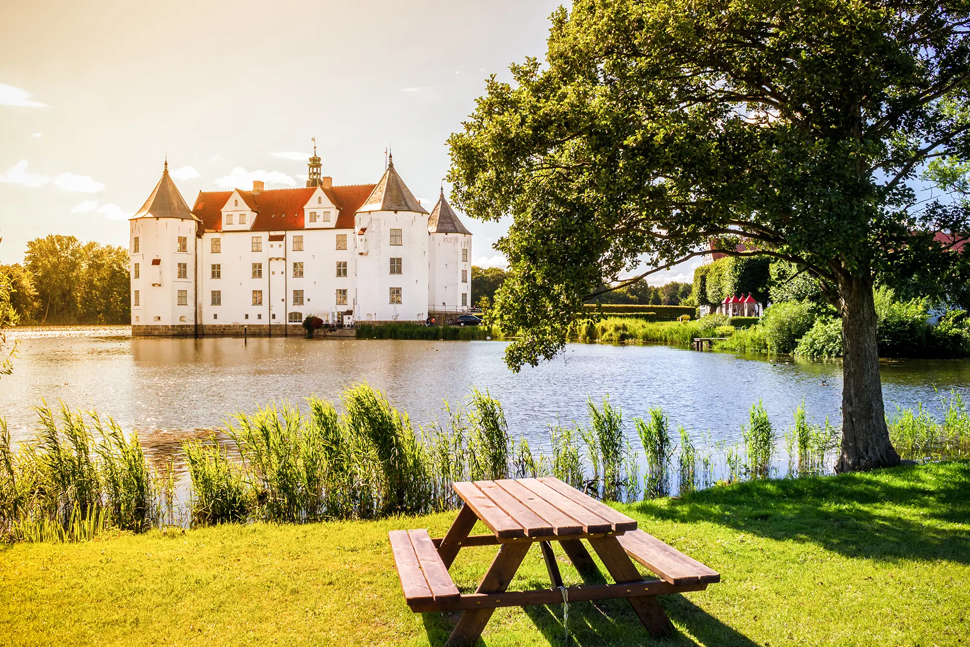 Schloss Glücksburg in Schleswig-Holstein, gesehen über den Schlossteich im Sommer, mit einem Baum und einer Bank im Vordergrund.