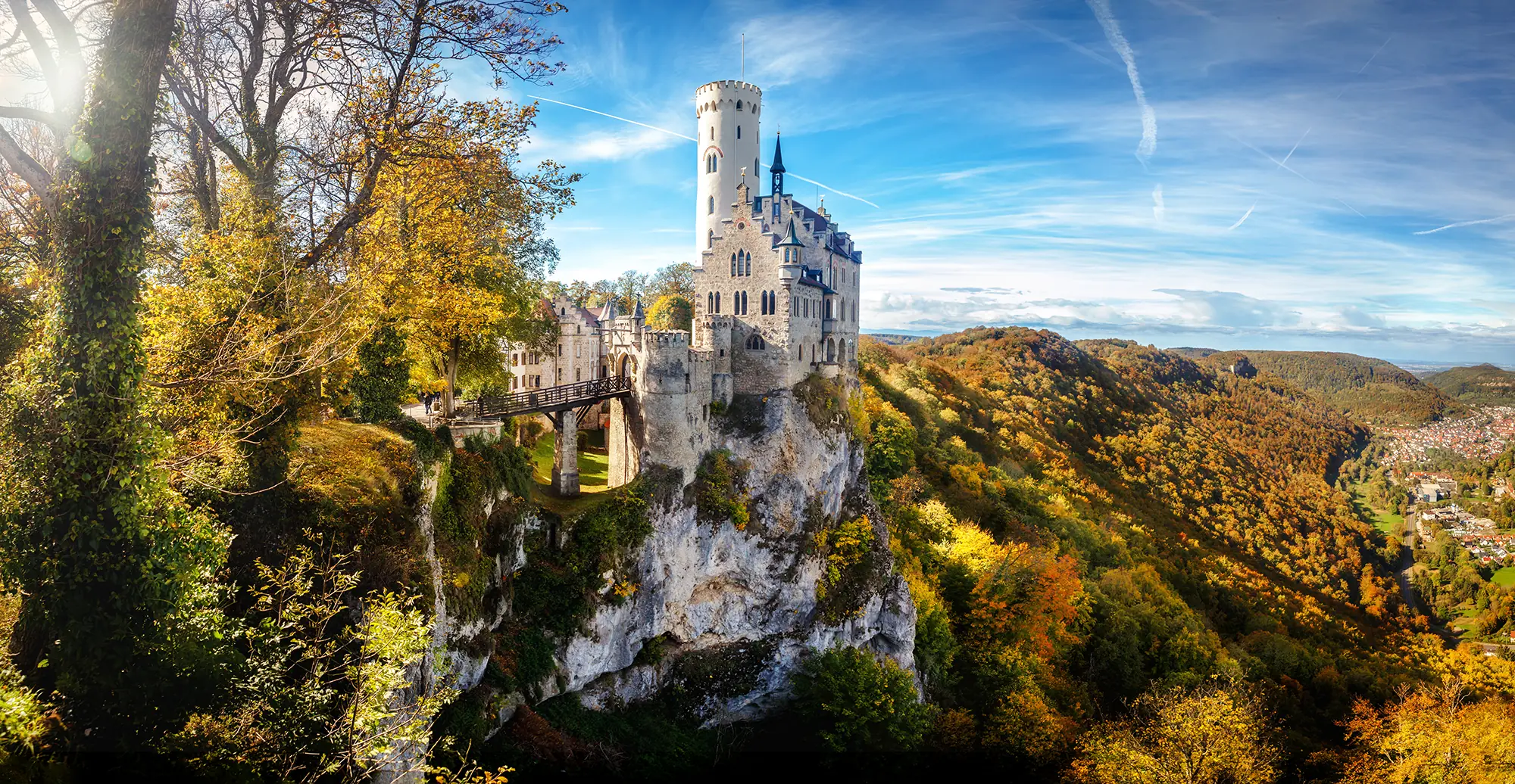 Panoramablick auf Schloss Lichtenstein mit der Brücke zum Schloss, umgeben von Bergen und Wäldern mit farbigem Laub im Hintergrund.