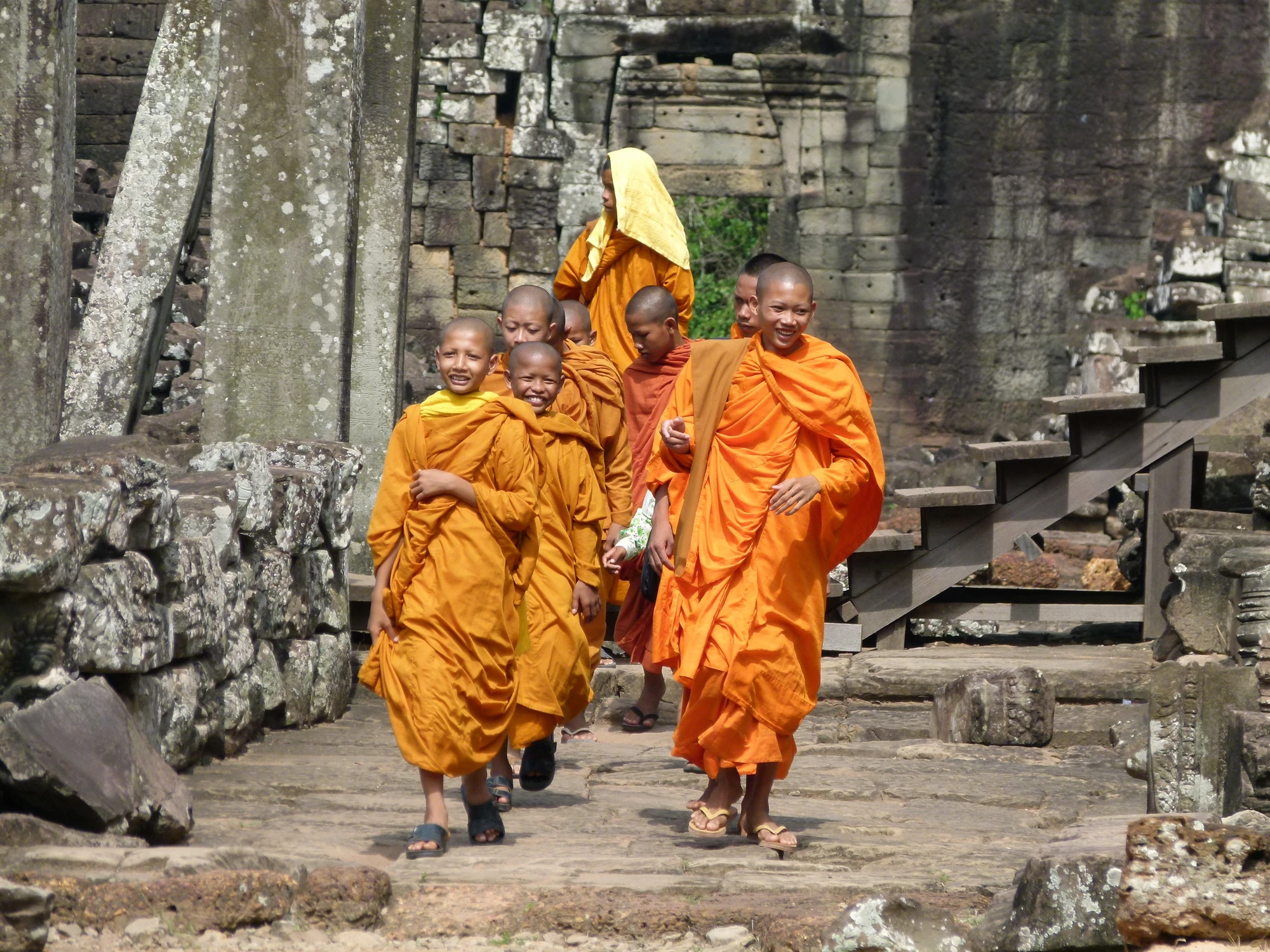 Fünf buddhistische Mönche in orangefarbenen Roben gehen auf einem steinigen Weg in einer Tempelanlage. Im Hintergrund sind alte Steine und Treppen sichtbar.