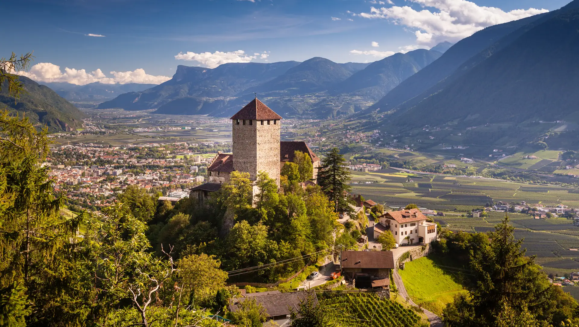 Burg Tirol in der Nähe von Bozen auf einem bewaldeten Hügel mit Tal und Bergen im Hintergrund unter blauem Himmel mit Wolken.