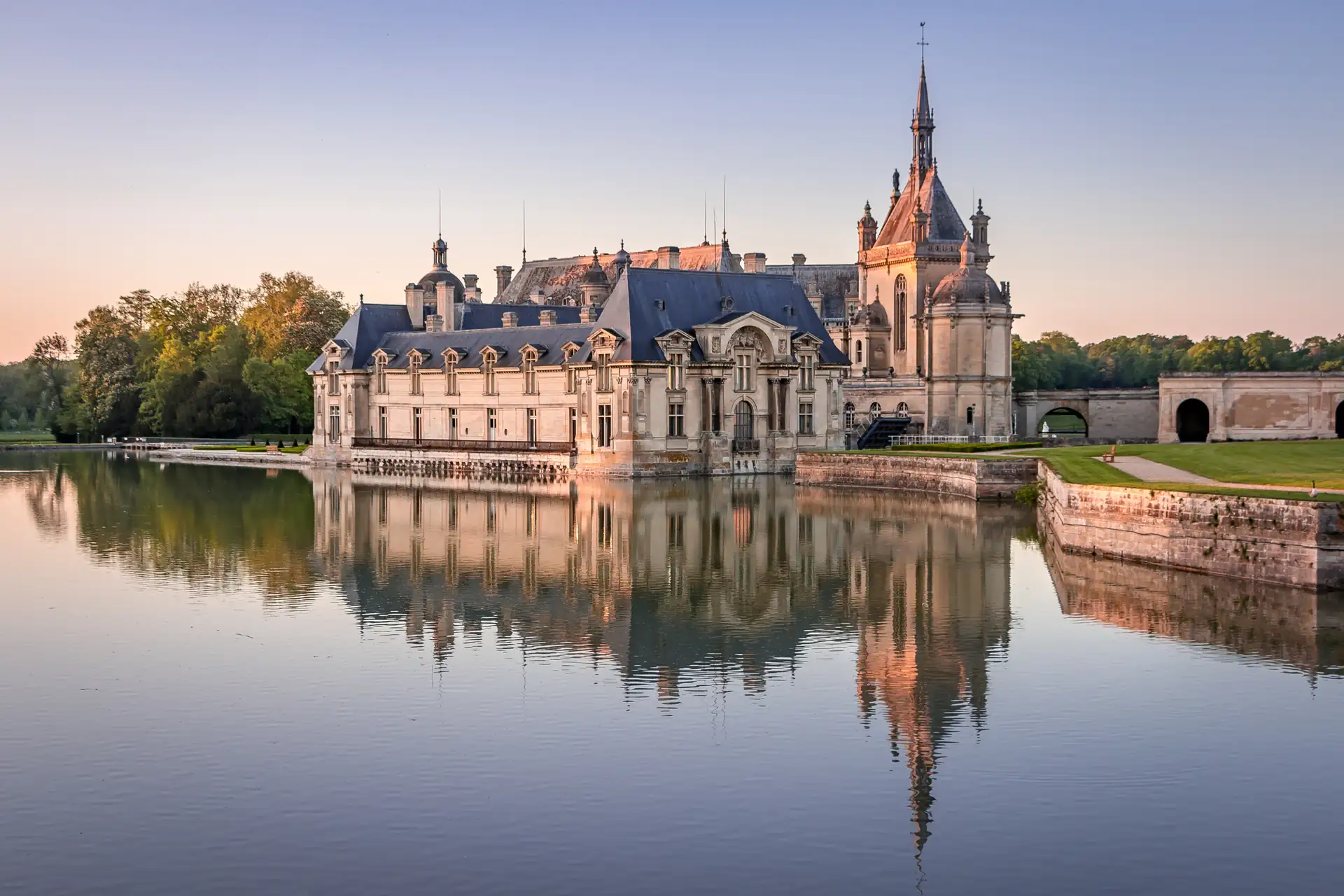 Château Chantilly mit Turm und Zinnen am Ufer eines ruhigen Gewässers, Spiegelung im Wasser, Bäume im Hintergrund.
