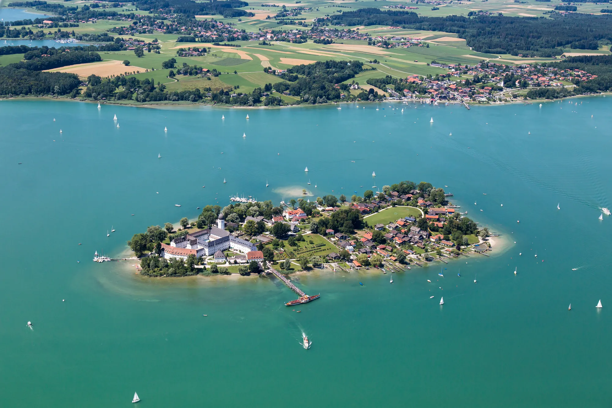 Das Bild zeigt eine Luftaufnahme der Fraueninsel im Chiemsee, Bayern, umgeben von türkisfarbenem Wasser. Die Insel ist dicht bebaut mit historischen Gebäuden, darunter das Kloster Frauenwörth, und sie ist von grünen Bäumen umgeben. Im Vordergrund sieht man die Anlegestelle, von der aus Boote zur Insel fahren, während Segelboote friedlich über den See gleiten. Im Hintergrund erstreckt sich das Festland mit Feldern und Dörfern. Das Gesamtbild vermittelt eine ruhige, malerische Landschaft mit einem charmanten Mix aus Natur und Kultur.