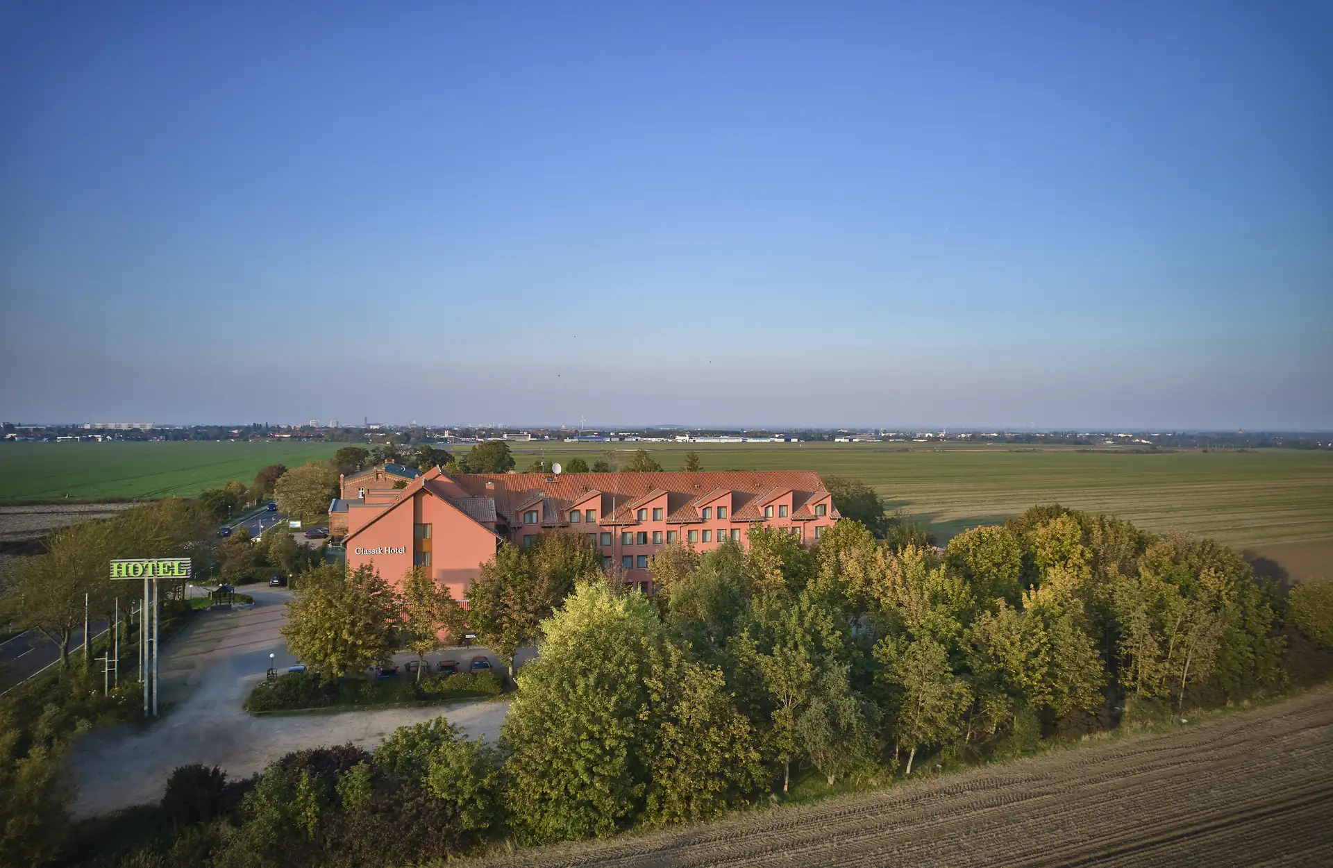 Das Classik Hotel Magdeburg als mehrstöckiges Gebäude mit rotem Dach umgeben von Bäumen und Feldern unter klarem Himmel