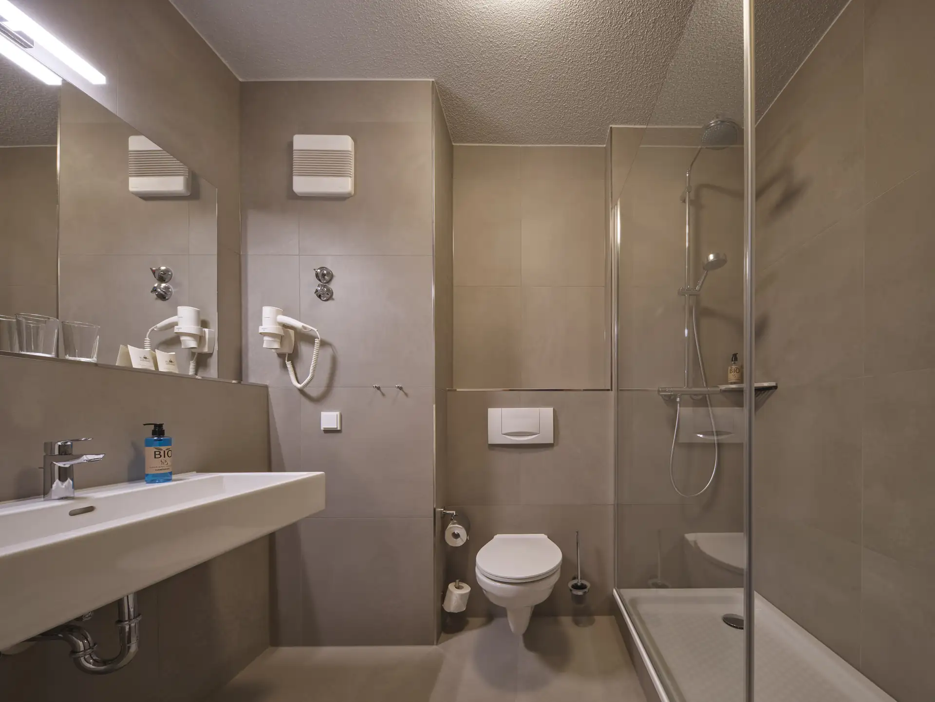 Modernes Hotelbad mit Wand-WC, großer Spiegelwand, Waschbecken mit Seifenspender, Haartrockner und Duschkabine mit Glasabtrennung