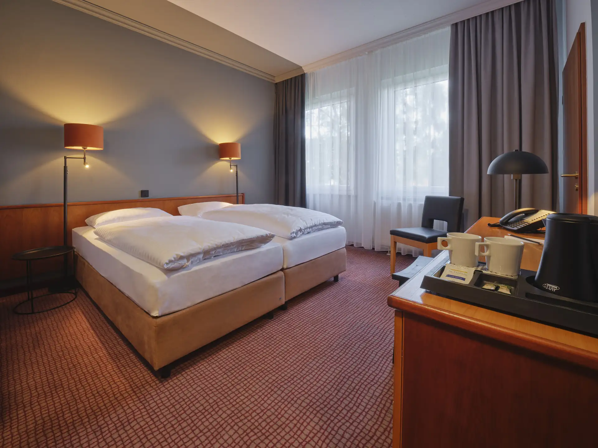 Komfortables Hotelzimmer mit zwei Einzelbetten, Nachttischlampen, Sessel vor Fenster mit Vorhängen und einem Tisch mit Teekanne und Tassen