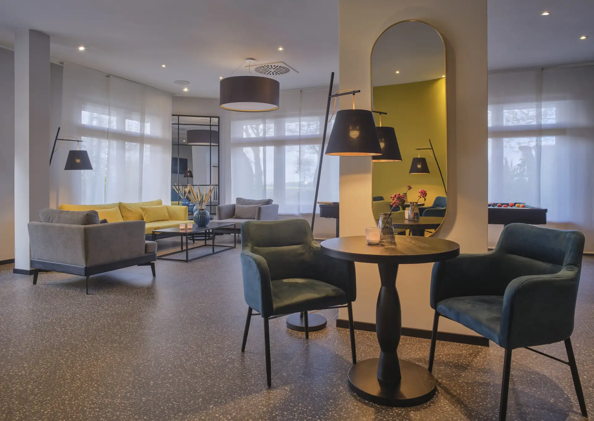 Moderne Hotellobby mit Sitzgruppen aus Sesseln und Sofas, Stehlampen, großem Wandspiegel und großen Fenstern mit weißen Vorhängen