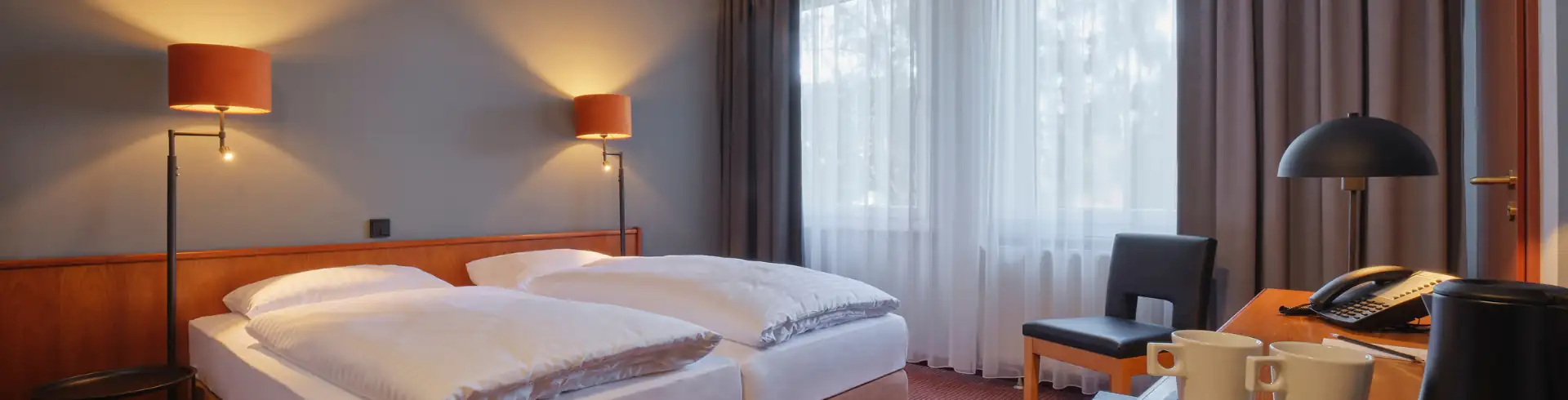 Komfortables Hotelzimmer mit zwei Einzelbetten, Nachttischlampen, Sessel vor Fenster mit Vorhängen und einem Tisch mit Teekanne und Tassen (Bannerformat)