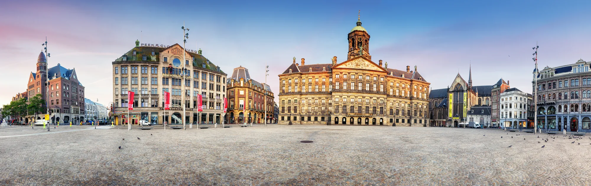 Panorama vom Dam Platz in Amsterdam, das das Madame Tussauds Museum und den Königspalast zeigt, umgeben von einer sanften Abenddämmerung und einigen Tauben, die auf dem Platz verweilen.