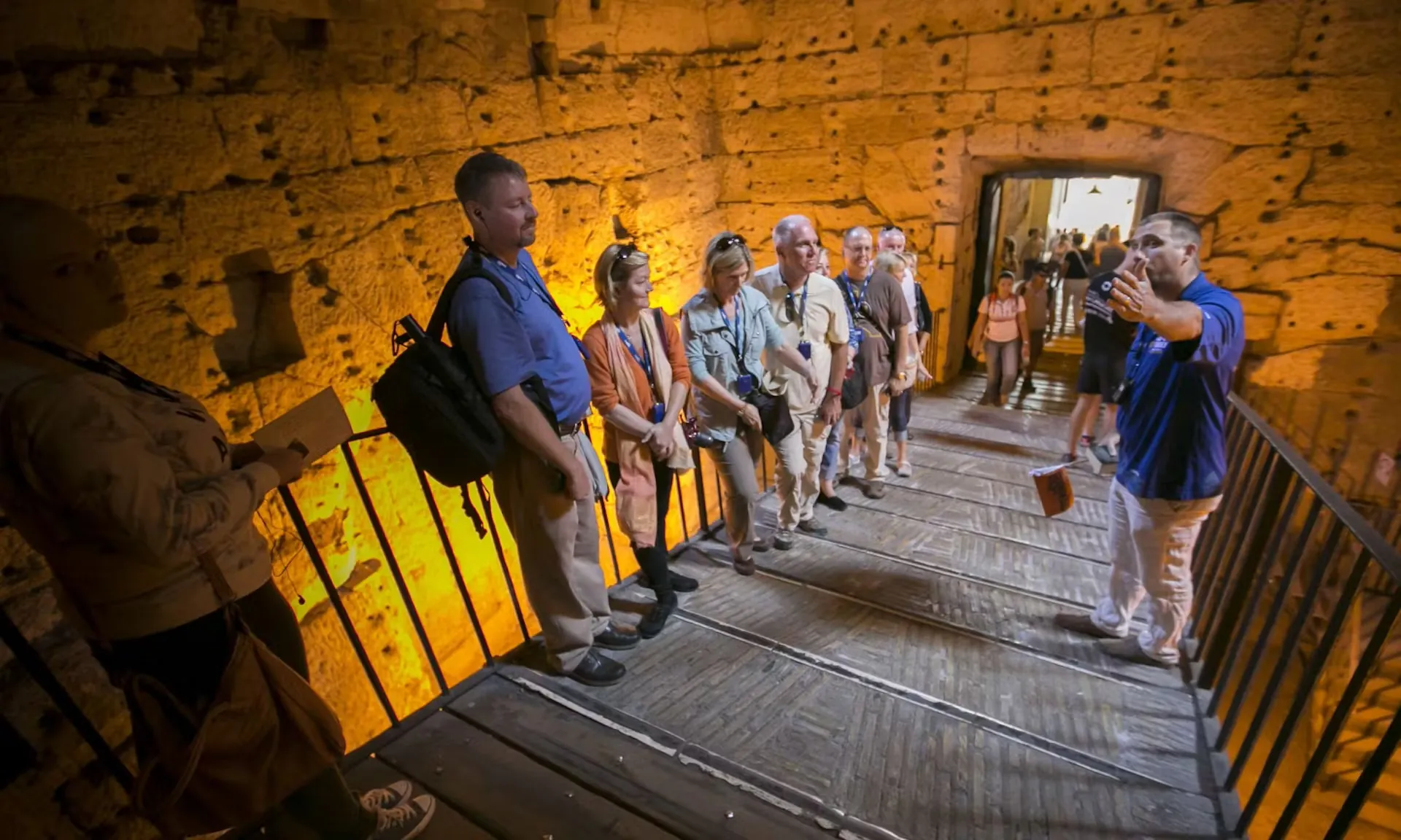 Das Bild zeigt eine Gruppe von Touristen, die an der Dan Brown Illuminati Tour in Rom teilnehmen. Ein Guide erklärt den Teilnehmern in einer historischen Kulisse, umgeben von massiven Steinwänden, die Geheimnisse der Stadt und ihrer Verbindung zu Dan Browns Romanen. Die Touristen hören aufmerksam zu, während der Guide gestikuliert, um seine Erklärungen zu untermalen. Die warme Beleuchtung verstärkt die geheimnisvolle Atmosphäre.