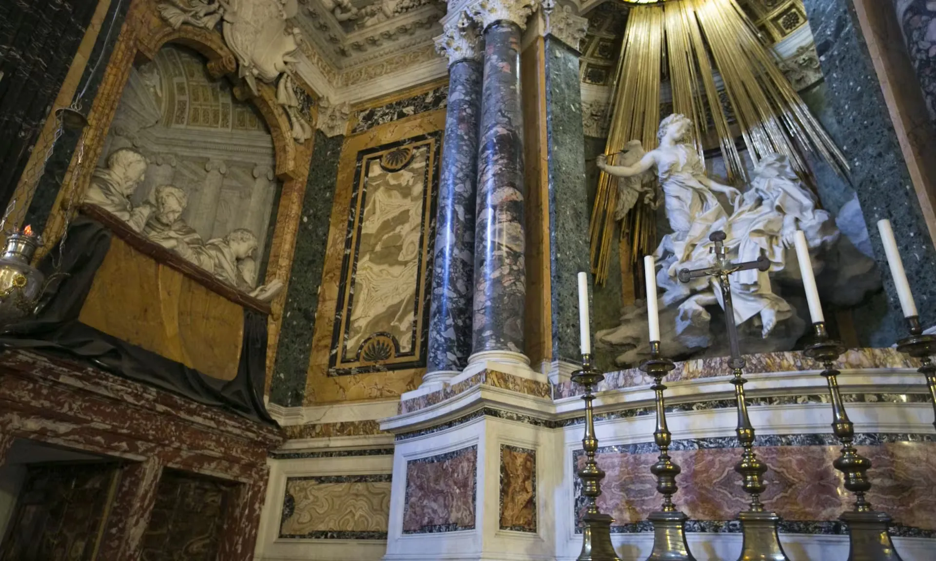 Das Bild zeigt ein kunstvoll gestaltetes Detail eines religiösen Altars, das Teil der Dan Brown Illuminati Tour ist. Im Fokus stehen mächtige Marmorsäulen, goldene Strahlen, die von oben herabfallen, und eine dramatische Skulptur einer heiligen Figur, vermutlich eines Engels oder einer Heiligen. Das Arrangement wird durch Kerzenleuchter und filigrane Verzierungen ergänzt. Im Hintergrund sind zudem Reliefs und aufwendige Mosaiken zu sehen. Diese Szene verkörpert die Pracht und den spirituellen Reichtum der besuchten Kirchen und Orte auf der Tour.