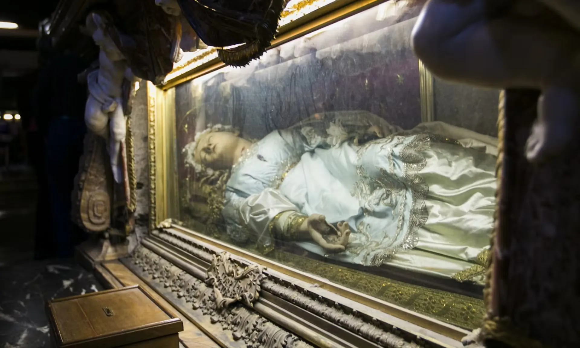 Das Bild zeigt eine kunstvoll gestaltete Glasvitrine, in der eine Puppe oder Figur in detailreich verzierter Kleidung liegt. Die Puppe stellt vermutlich eine religiöse Figur dar und befindet sich in einem prunkvollen, vergoldeten Rahmen. Diese Szene stammt von der Dan Brown 