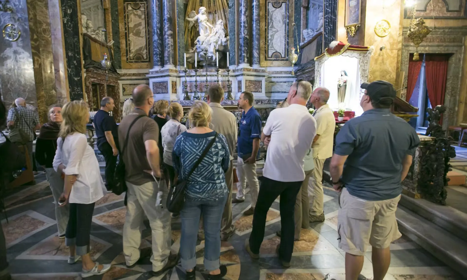 Das Bild zeigt eine Gruppe von Touristen, die an der Dan Brown „Illuminati“-Tour teilnehmen. Sie stehen in einer reich verzierten Kirche und betrachten aufmerksam die kunstvolle Marmorskulptur „Die Verzückung der Heiligen Theresa“ von Gian Lorenzo Bernini. Das prachtvolle Interieur der Kirche mit seinen Marmorsäulen und goldenen Akzenten verleiht der Szene eine ehrwürdige Atmosphäre. Die Touristen scheinen gespannt den Erklärungen des Führers zu lauschen, während sie sich in dieser historisch und künstlerisch bedeutsamen Umgebung aufhalten.