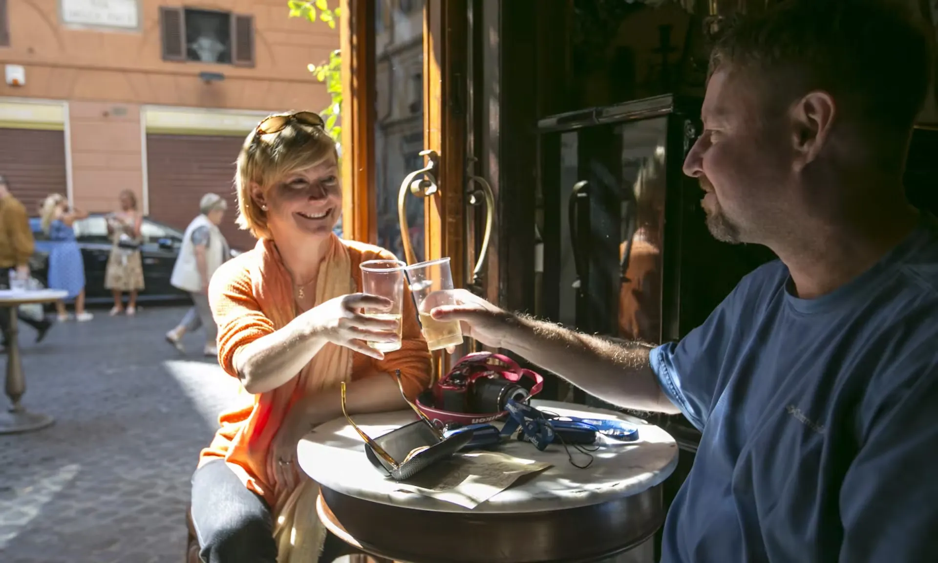 Das Bild zeigt zwei Personen, die während einer Pause auf der Dan Brown „Illuminati“-Tour in einem gemütlichen Café anstoßen. Sie sitzen an einem kleinen runden Tisch in entspannter Atmosphäre, die Sonnenstrahlen fallen durch die Tür und erhellen den Raum. Im Hintergrund sind unscharf weitere Touristen auf einer gepflasterten Straße zu sehen. Die Szene strahlt Gelassenheit aus und spiegelt den kulturellen Genuss dieser literarischen Tour wider.