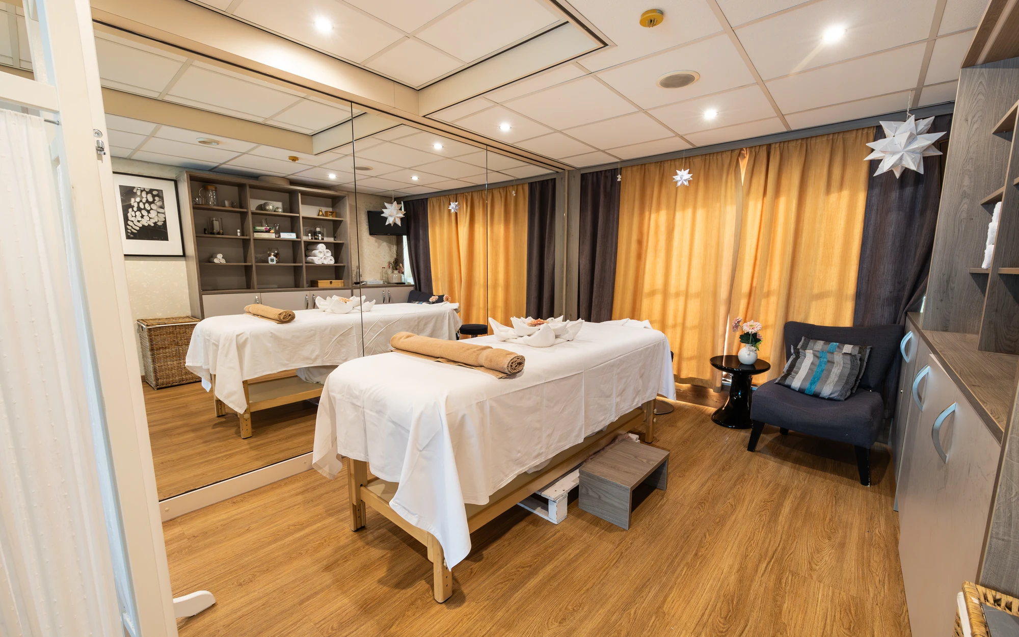 Massageraum mit Massageliege mit einer Blume aus Handtüchern gefaltet vor einem Spiegel und einem Sessel und Schränken