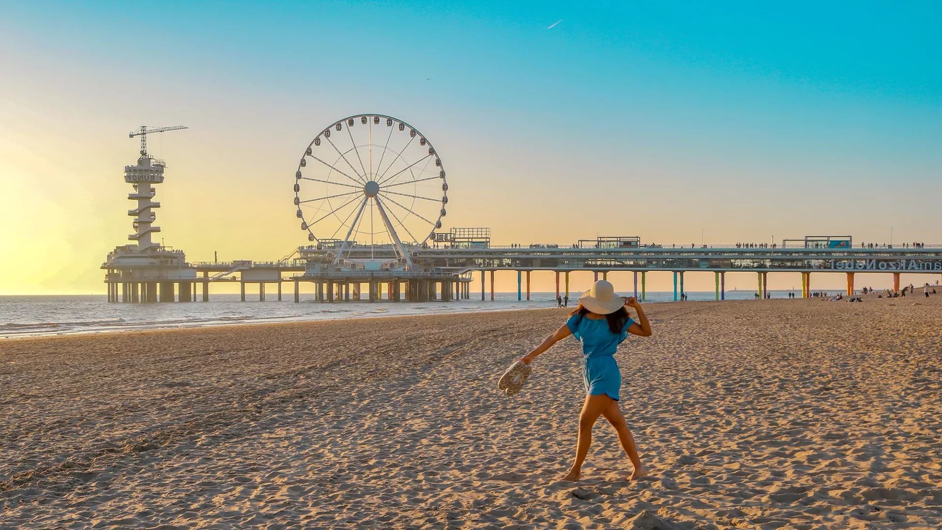 Eine Frau im kurzen blauen Overall und Strohhut spaziert am Pier von Scheveningen entlang, während die sanfte Dämmerung den Himmel in sanften Farben taucht. Hinter ihr erstreckt sich das Meer, dessen Wellen sanft gegen den Strand schlagen, und das ikonische Riesenrad fügt der Szenerie einen charmanten Akzent hinzu. Die ruhige Atmosphäre und das abendliche Licht schaffen eine malerische Kulisse für diesen entspannten Strandspaziergang.