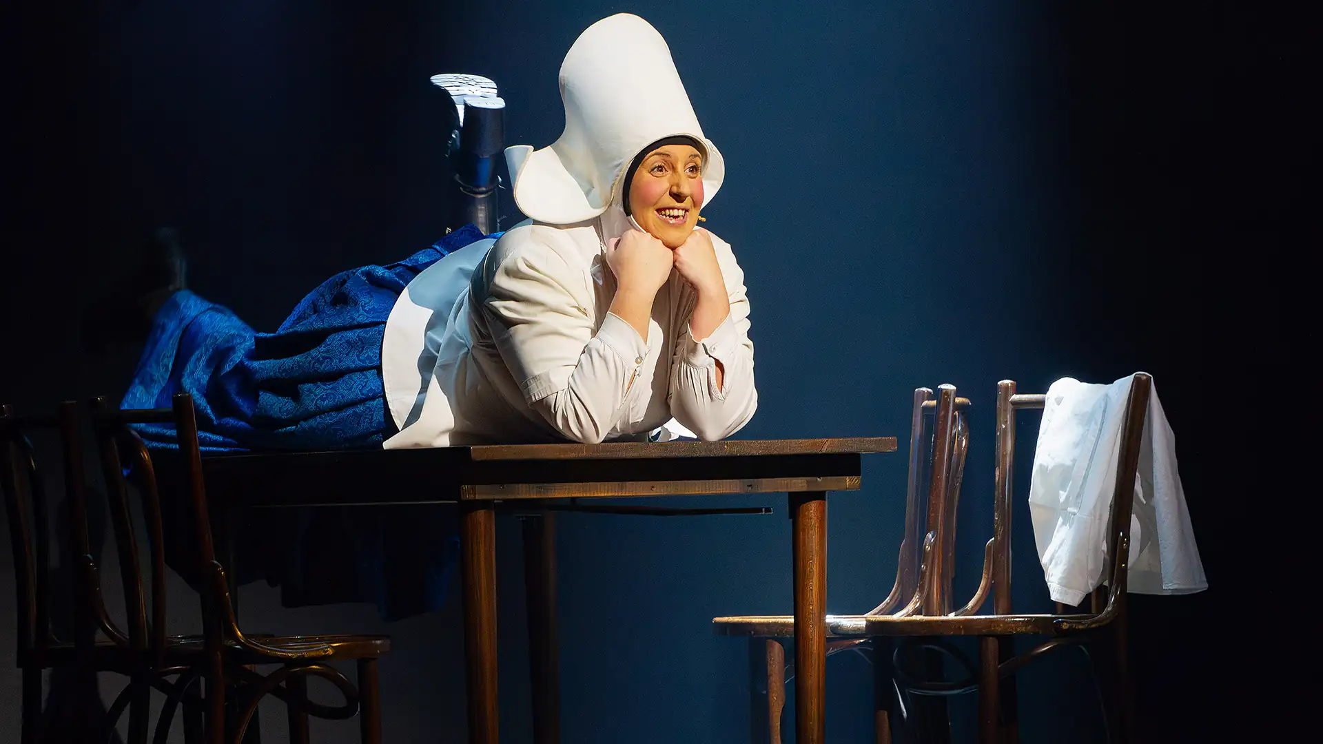 Die AMME - Das Musical Szenenbild (Kachel): Frau in weiß-blauer Kleidung mit Haube fröhlich auf einem Tisch liegend, daneben ein Stuhl.