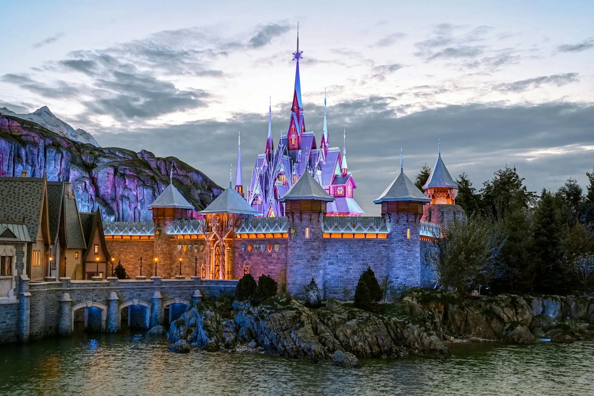 Das beleuchtete Schloss von Arendelle im Disneyland Paris bei Dämmerung mit umliegenden Türmen, Wasser und Felsen im Vordergrund