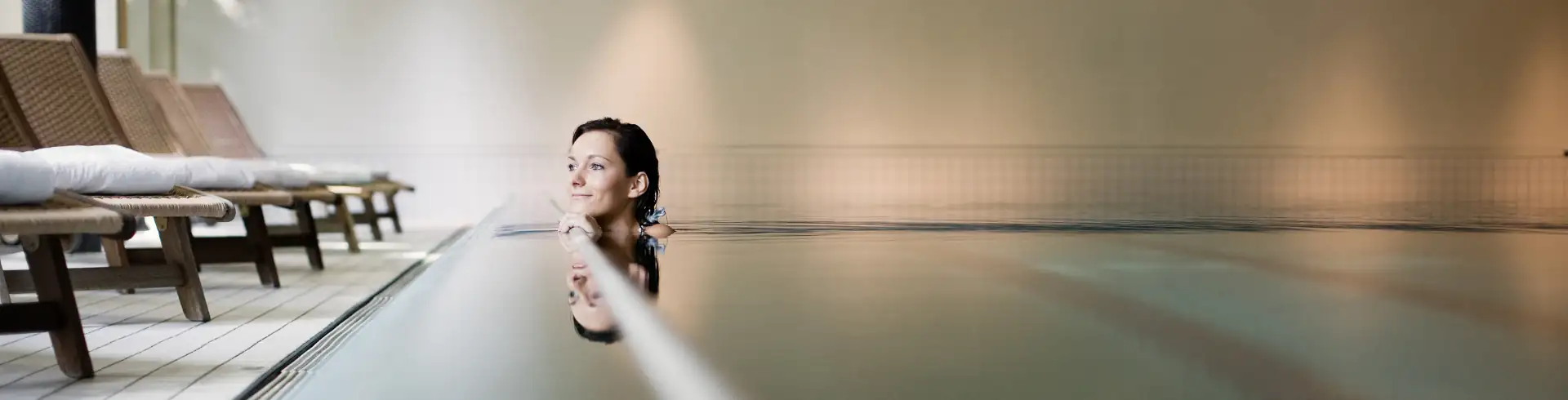 Dolce by Wyndham Bad Nauheim im Bannerformat: Frau entspannt im Hallenbad des Dolce Bad Nauheim mit Liegestühlen am Beckenrand.