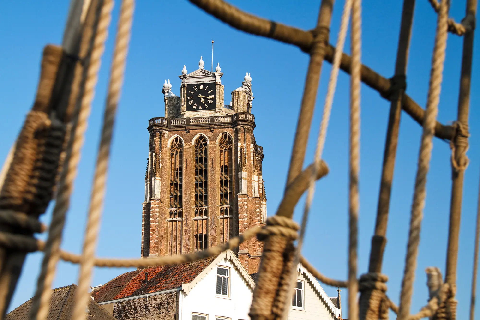 Eine Nahaufnahme des oberen Teils der Kathedrale in Dordrecht, eingefangen bei klarem Himmel. Die filigranen Details der gotischen Architektur und die beeindruckenden Türme sind deutlich sichtbar und heben die kunstvolle Gestaltung des Bauwerks hervor. Der strahlend blaue Himmel bietet einen schönen Kontrast und verstärkt die majestätische Ausstrahlung der Kathedrale.