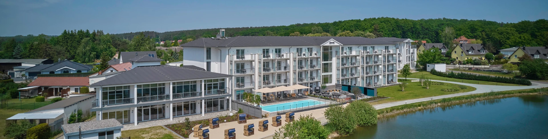 Ansicht eines Dorint Hotels mit mehreren Etagen, großen Fenstern und Balkonen, umgeben von Rasen und einem Teich. Im Vordergrund liegen Liegen am Pool.