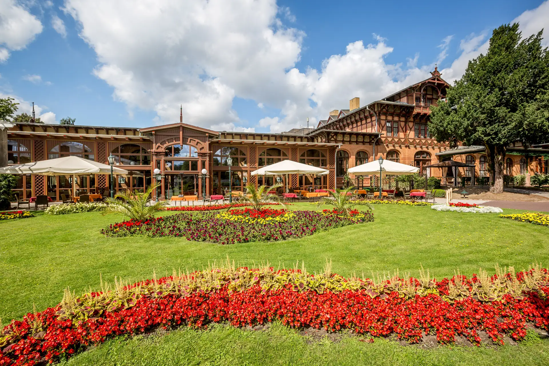 Das Dorint Herrenkrug Parkhotel Magdeburg mit großer Glasfassade, davor ein gepflegter Garten mit bunten Blumenbeeten und Sonnenschirmen auf der Terrasse