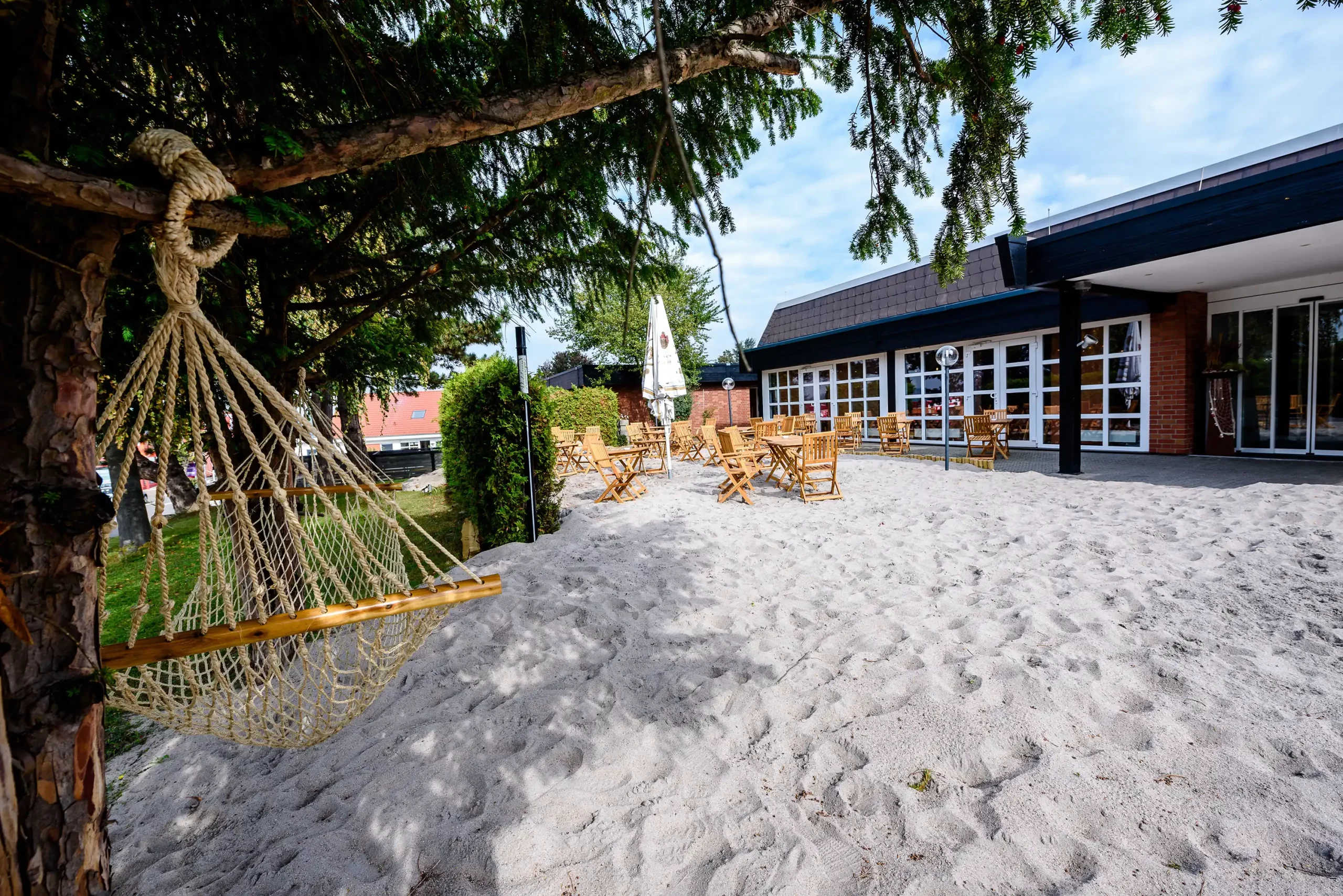 Außenbereich des Dorint Hotels Alzey/Worms mit Sandboden, Holzstühlen und Tischen sowie einer Hängematte an einem Baum