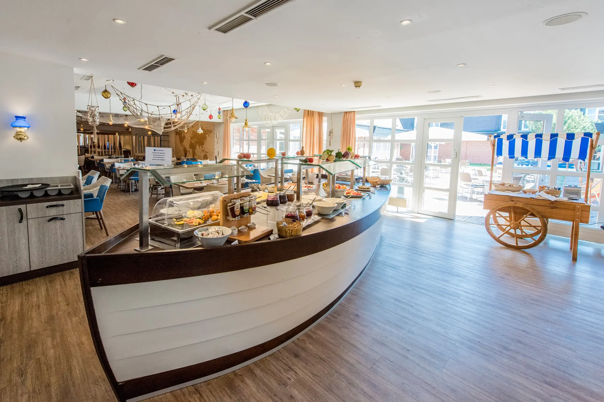 Halbrunder Frühstücksbuffet-Tresen in Form eines Bootes mit verschiedenen Speisen in einem hellen Hotelrestaurant mit großen Fenstern und Holzboden