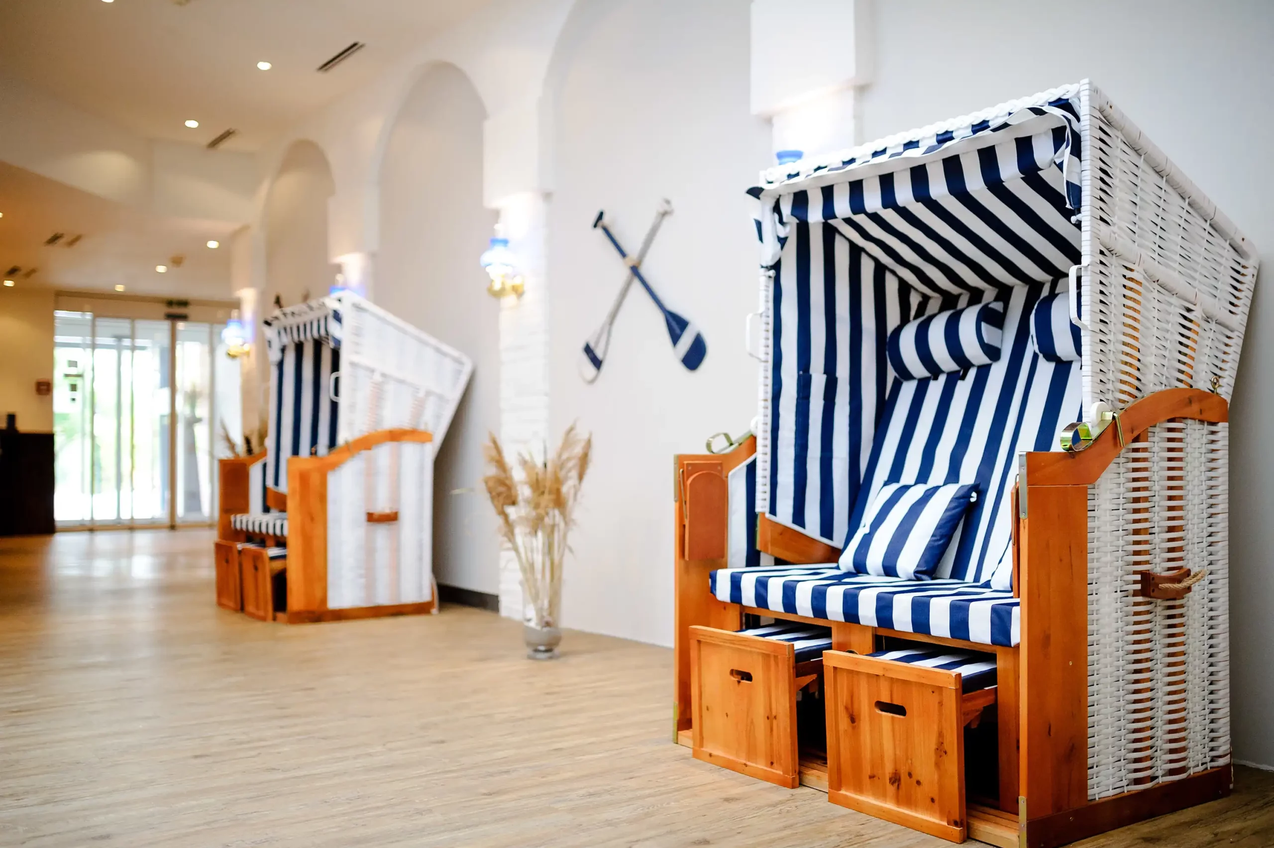Lobby des Dorint Hotels Alzey/Worms mit mehreren Strandkorb-Sesseln aus Holz und weiß-blau gestreiftem Stoff sowie zwei gekreuzten Paddeln an der Wand und einer Vase mit getrocknetem Pampasgras auf hellem Holzboden