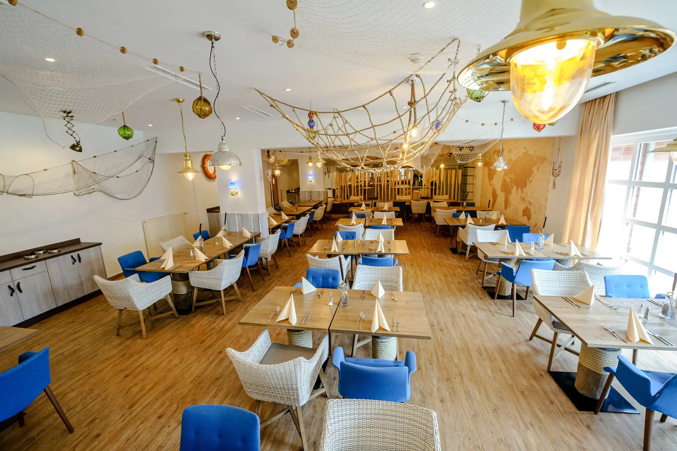 Weitwinkelaufnahme eines hellen Restaurants im Dorint Hotel Alzey/Worms mit Holzboden, blauen und weißen Stühlen, rechteckigen Tischen und maritimer Dekoration an der Decke