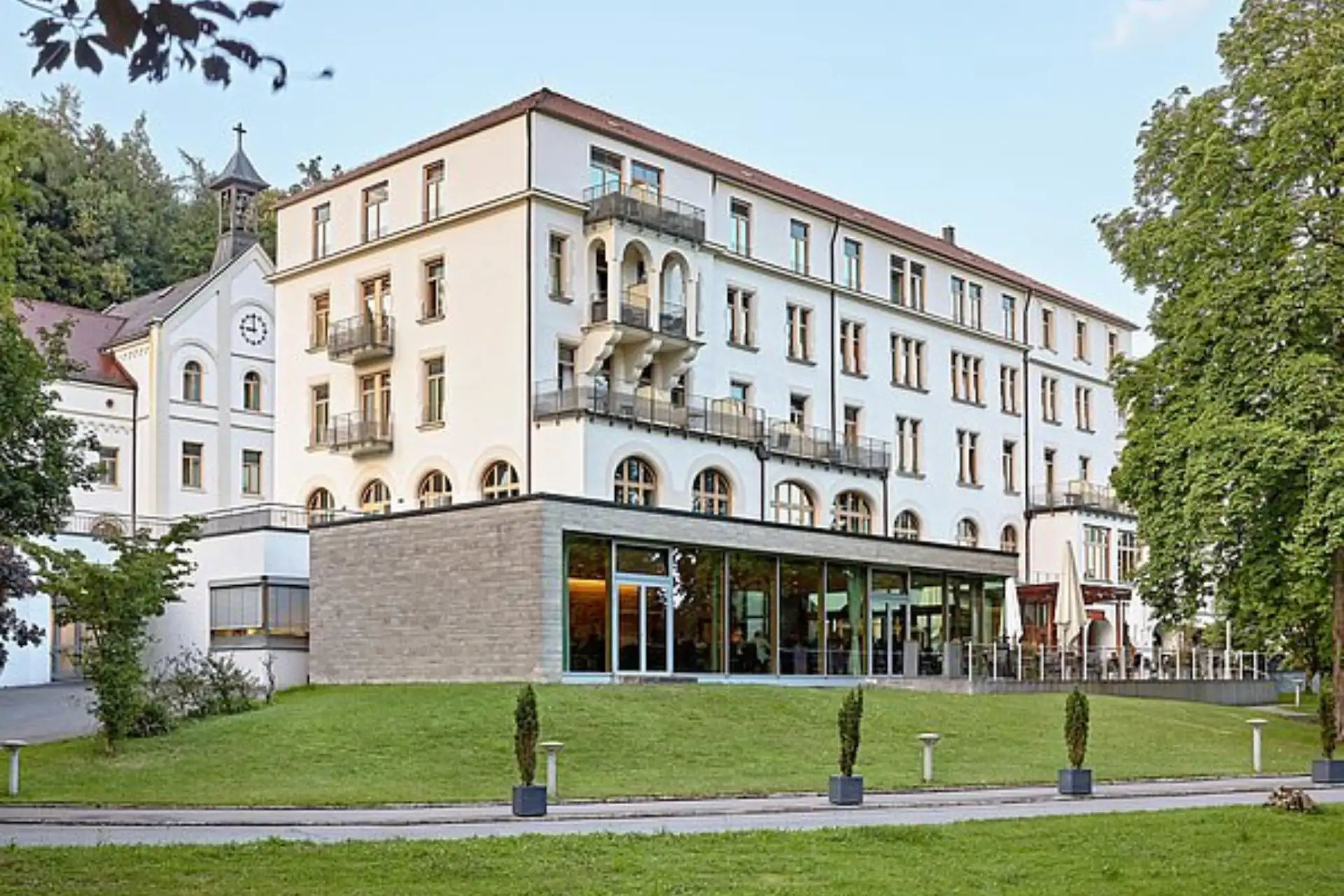Mehrstöckiges Hotelgebäude des Dorint Parkhotel Jordanbad Biberach an der Riß mit großem Glasanbau und angrenzender Kapelle vor bewaldetem Hintergrund