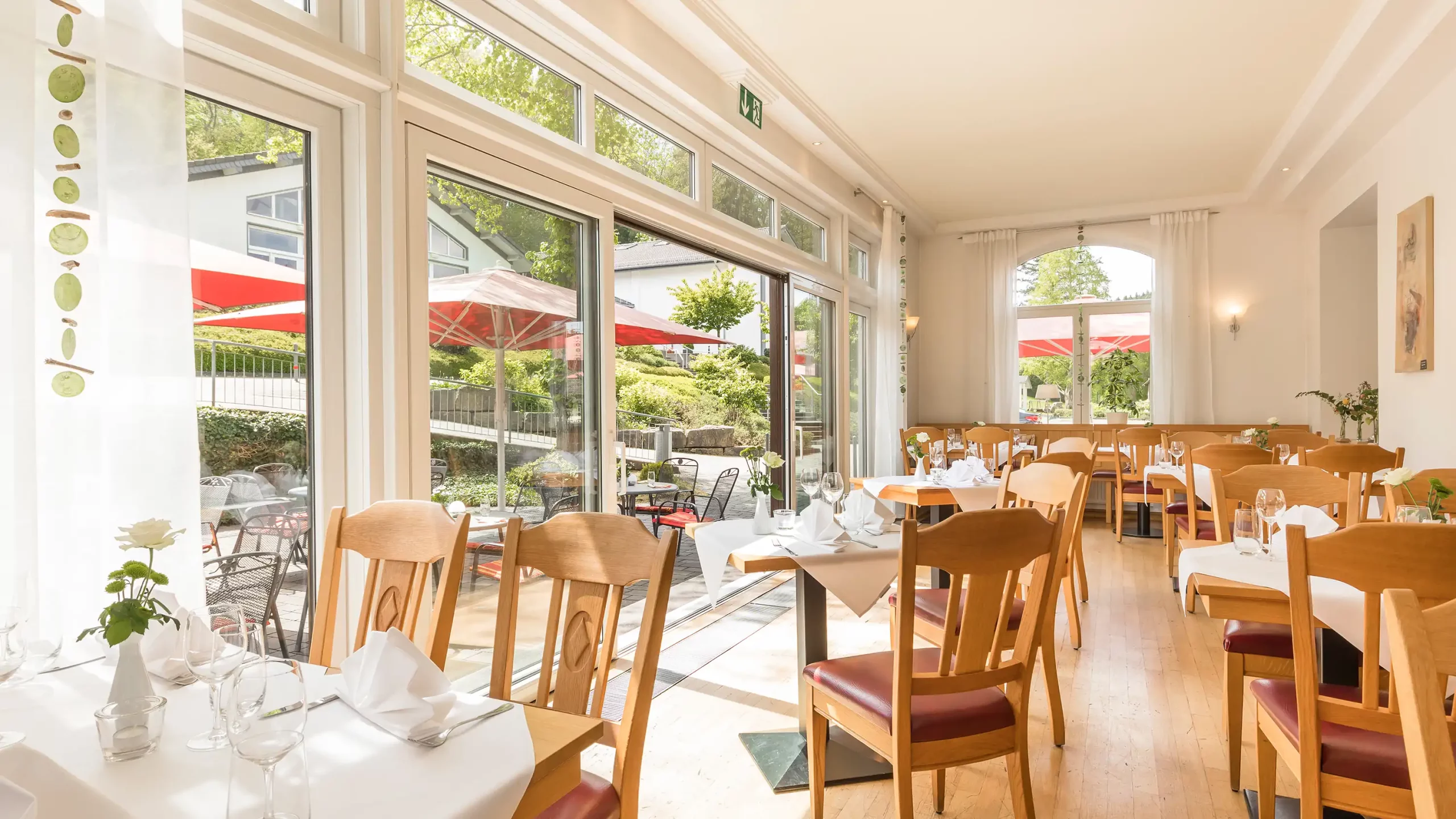 Helles Restaurant mit Holztischen und Stühlen, weiß gedeckten Tischen und großer Glasfront zur Terrasse mit roten Sonnenschirmen im Dorint Parkhotel Siegen
