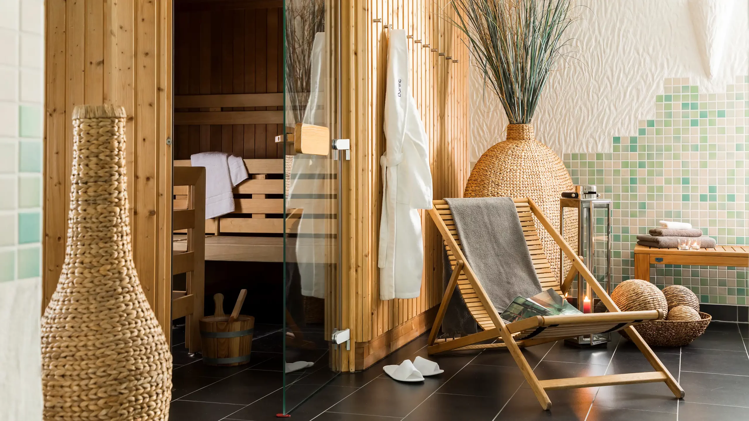 Saunabereich mit Holzbänken, Glas- und Holztür, geflochtener Vase, Liegestuhl mit grauem Handtuch und dekorativen Elementen auf dunklem Boden im Dorint Parkhotel Siegen