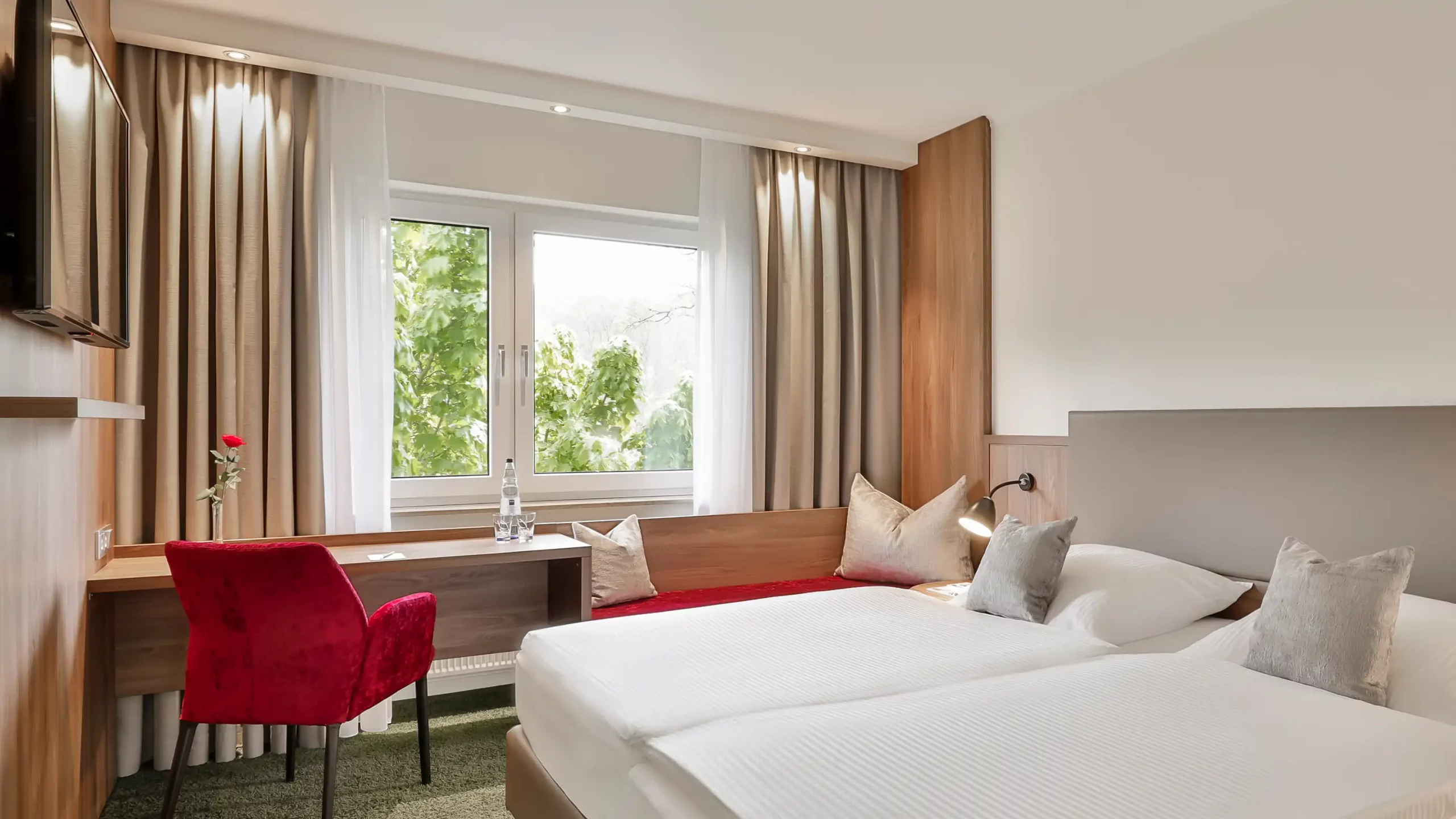 Modern eingerichtetes Superior-Zimmer mit Doppelbett, Schreibtisch, rotem Stuhl und großem Fenster mit Blick auf grüne Bäume im Dorint Parkhotel Siegen