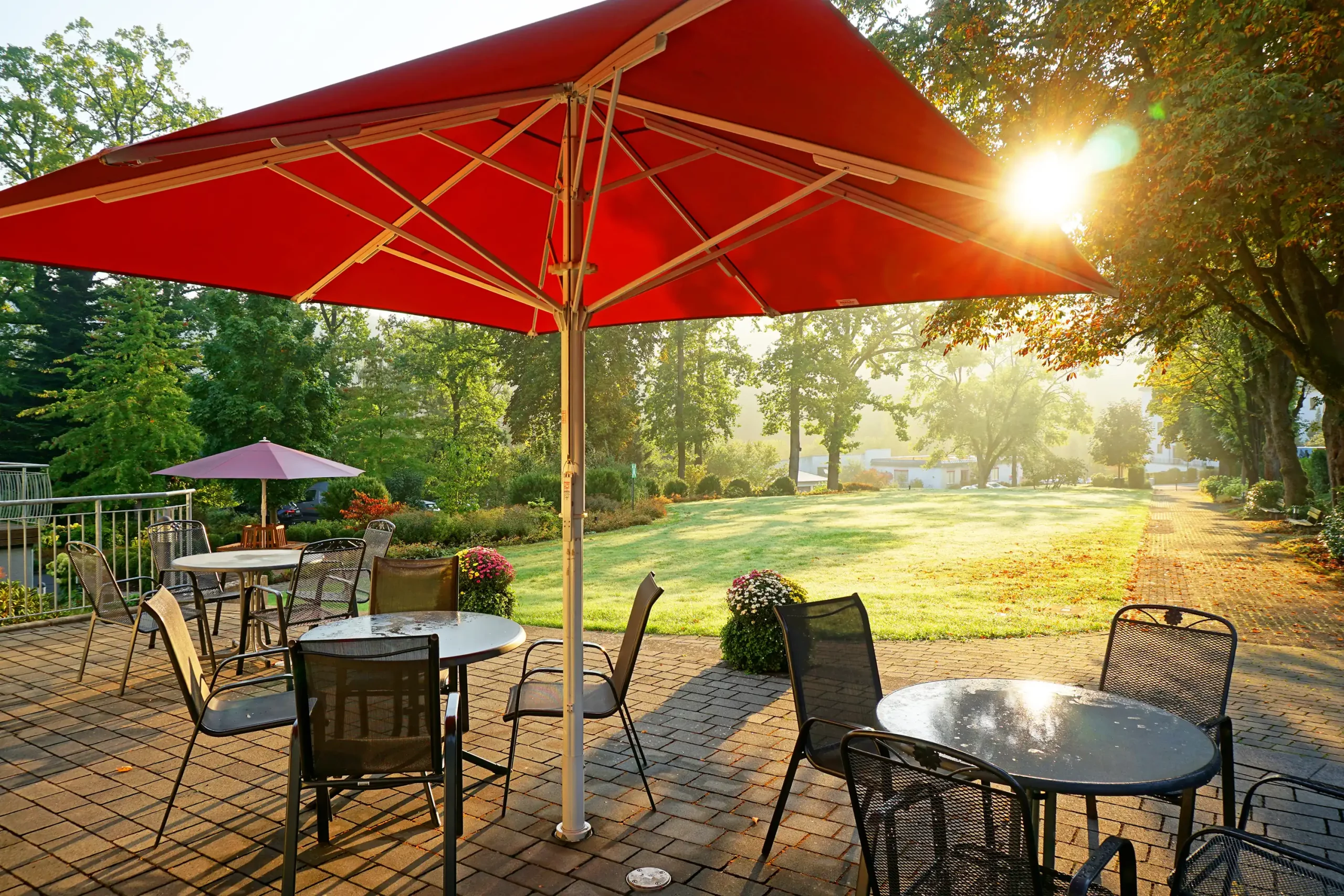 Terrasse mit Tischen, Stühlen und großen Sonnenschirmen auf gepflastertem Boden vor einer grünen Parklandschaft im Morgenlicht