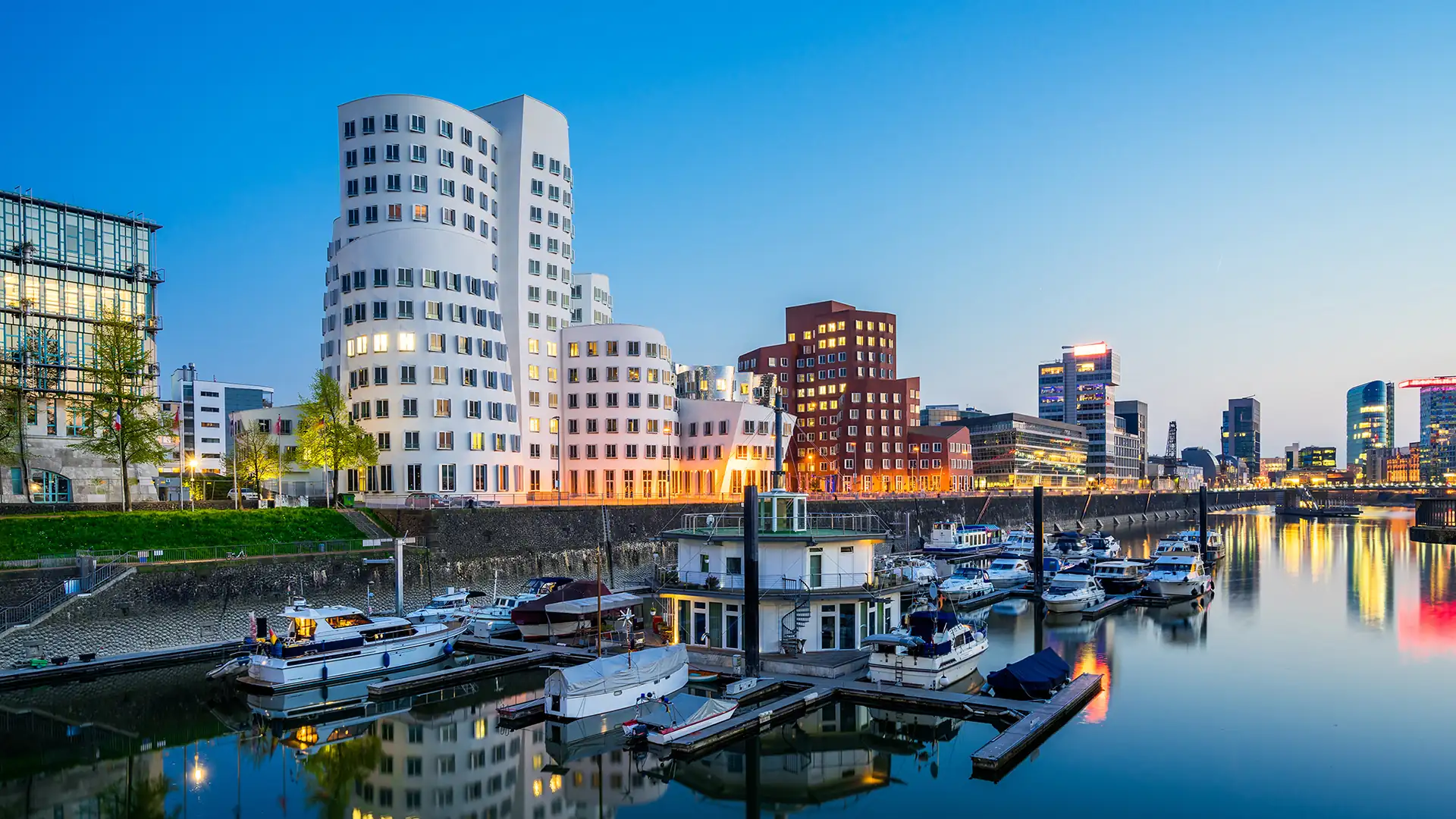 Beleuchtete Gebäude im Düsseldorfer Medienhafen zur Abenddämmerung mit Booten auf dem Rhein im Vordergrund (1920x1080px)