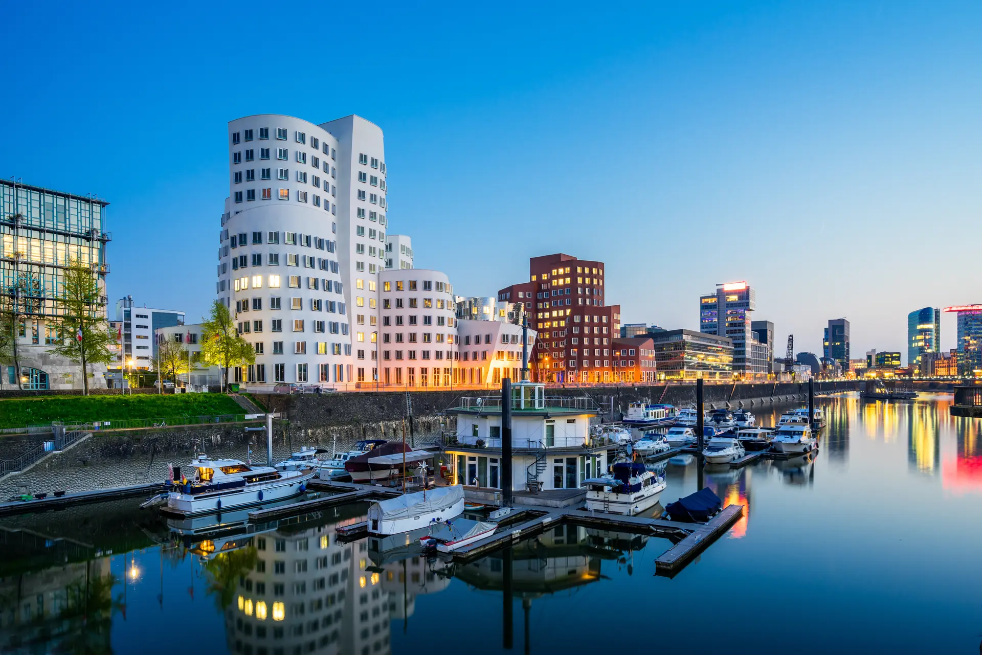Beleuchtete Gebäude im Düsseldorfer Medienhafen zur Abenddämmerung mit Booten auf dem Rhein im Vordergrund