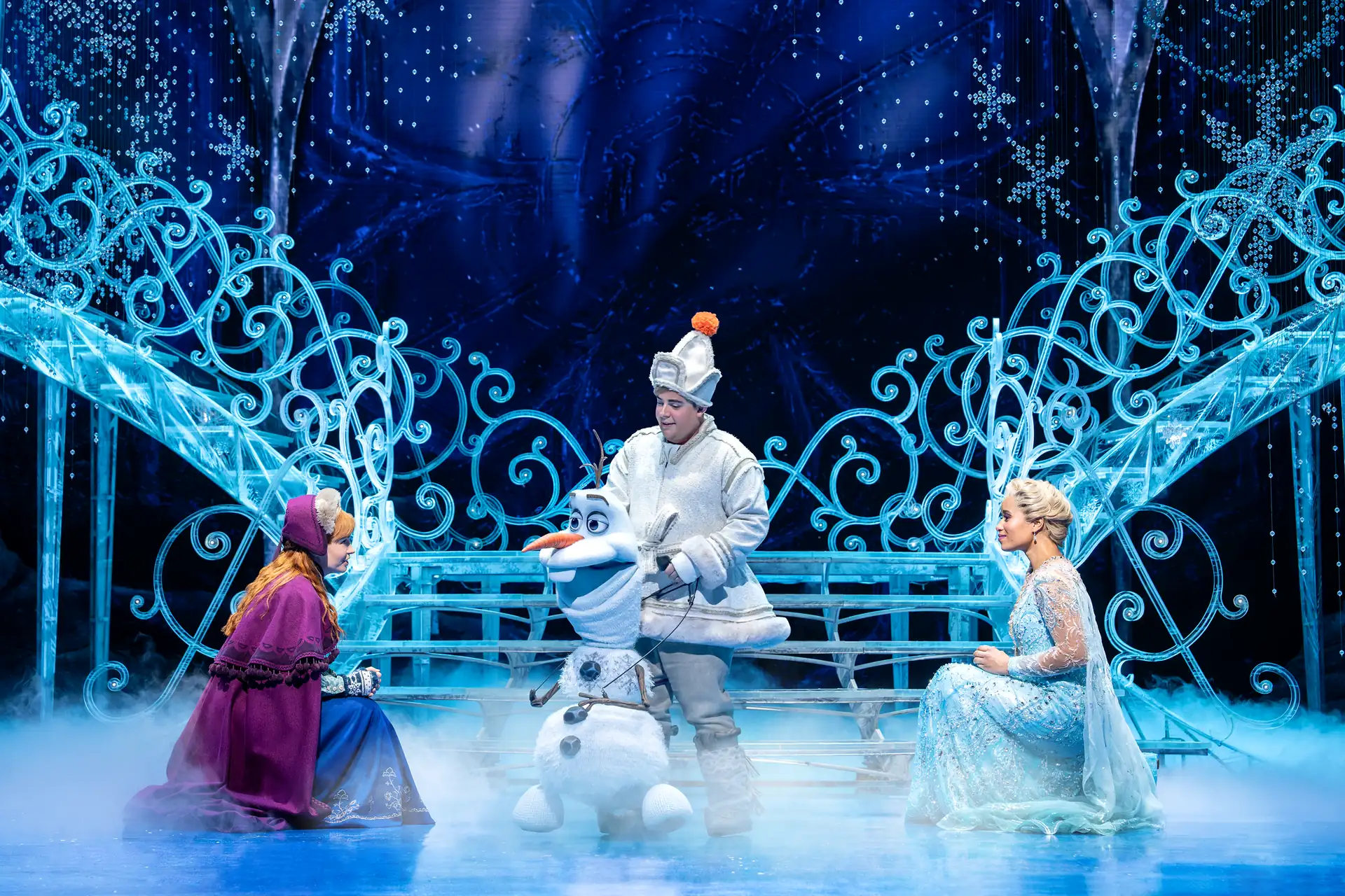 Drei Darsteller in Kostümen von Anna, Elsa und Olaf sitzen auf einer Bühne mit blauem, winterlichem Bühnenbild und Schneeflockendekorationen.